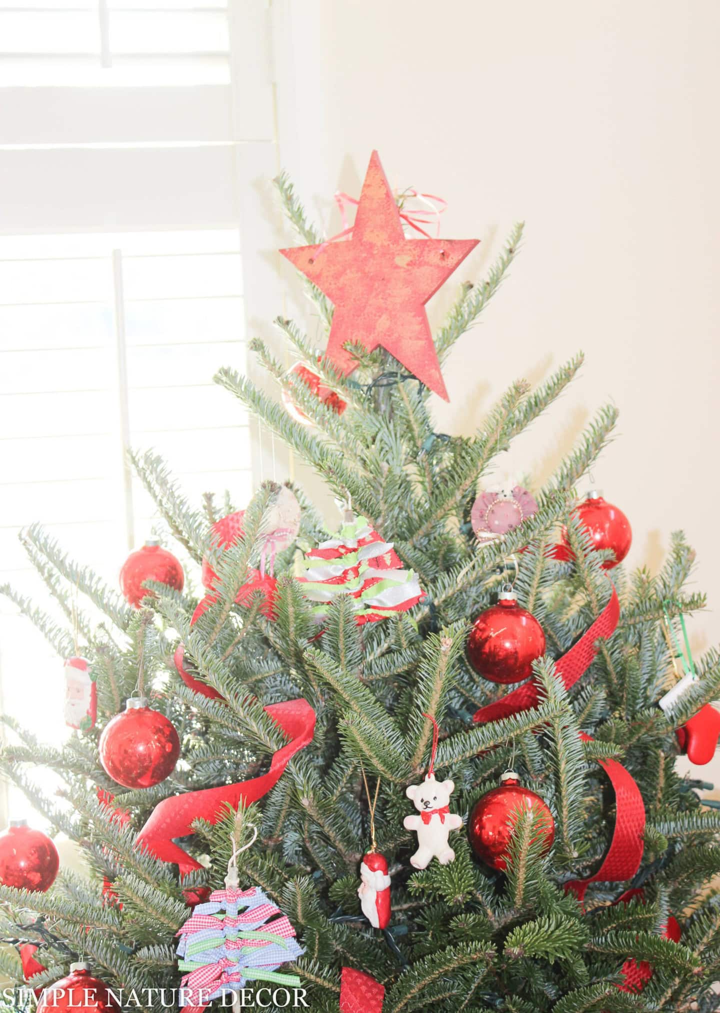A decorated Christmas tree with a red star topper, red ornaments, ribbon, and various festive hanging decorations near a window.
