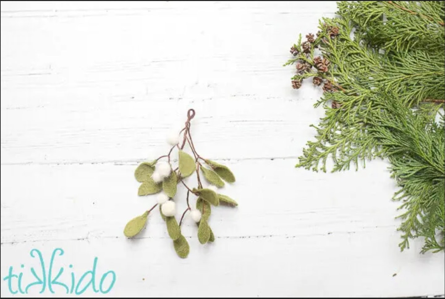 Felt mistletoe with white berries and green leaves lies next to a sprig of evergreen on a white wooden surface. The word