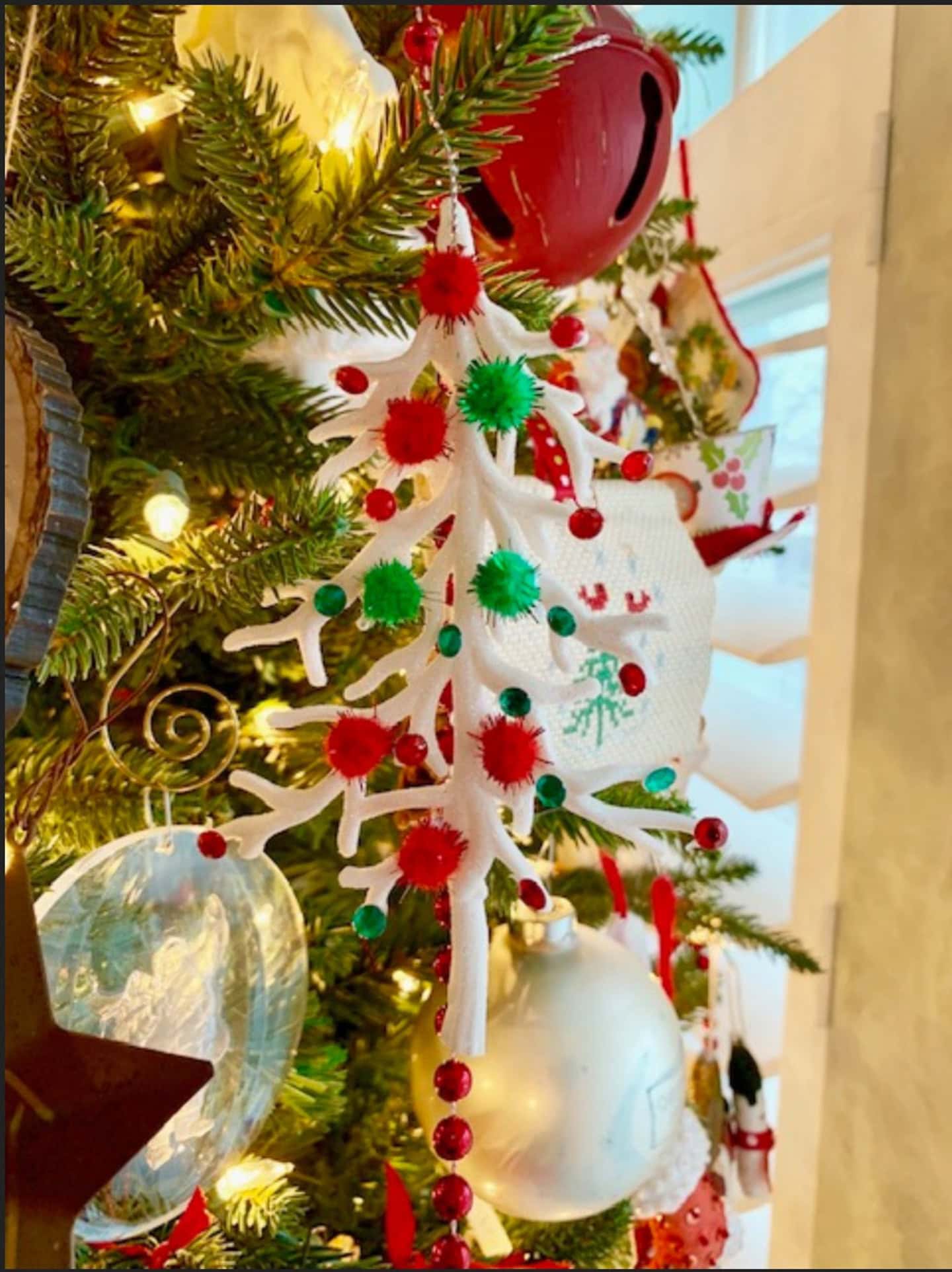 Close-up of a Christmas tree decorated with a white pipe cleaner ornament adorned with red and green pom-poms, red beads, and various festive ornaments in the background.