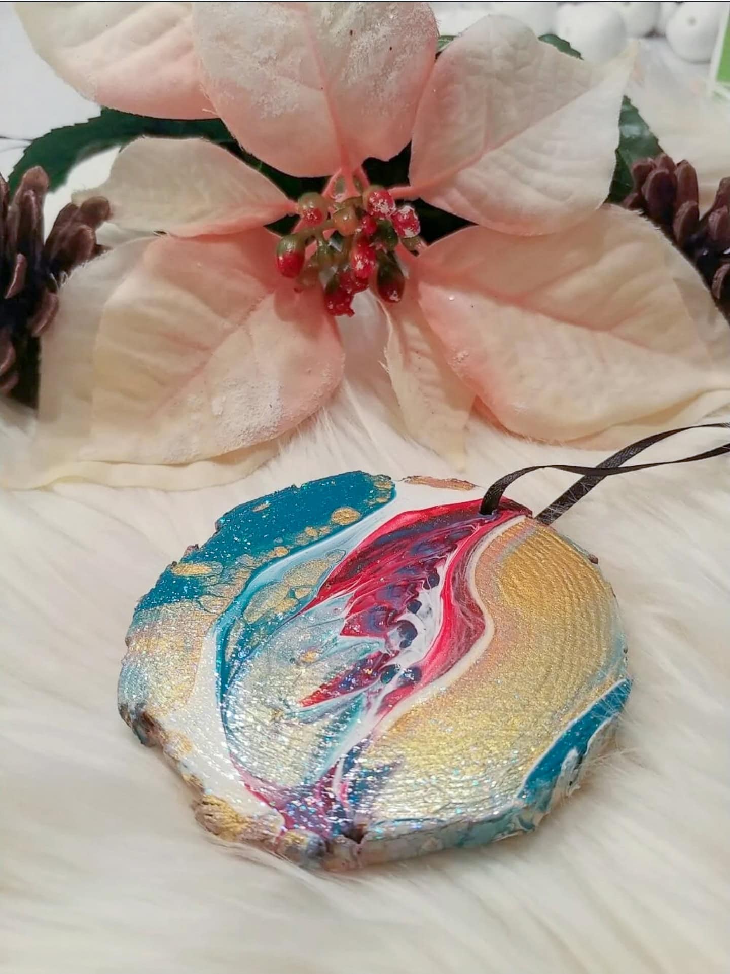A round, painted ornament with abstract swirls of blue, red, gold, and white rests on a white fur surface in front of a cream-colored artificial poinsettia flower.