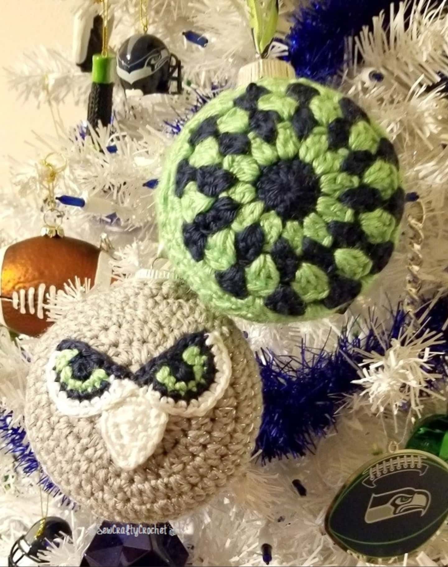 Two crocheted Christmas ornaments, one with an owl face and one with a green and blue circular pattern, hang on a white Christmas tree decorated with football-themed ornaments.