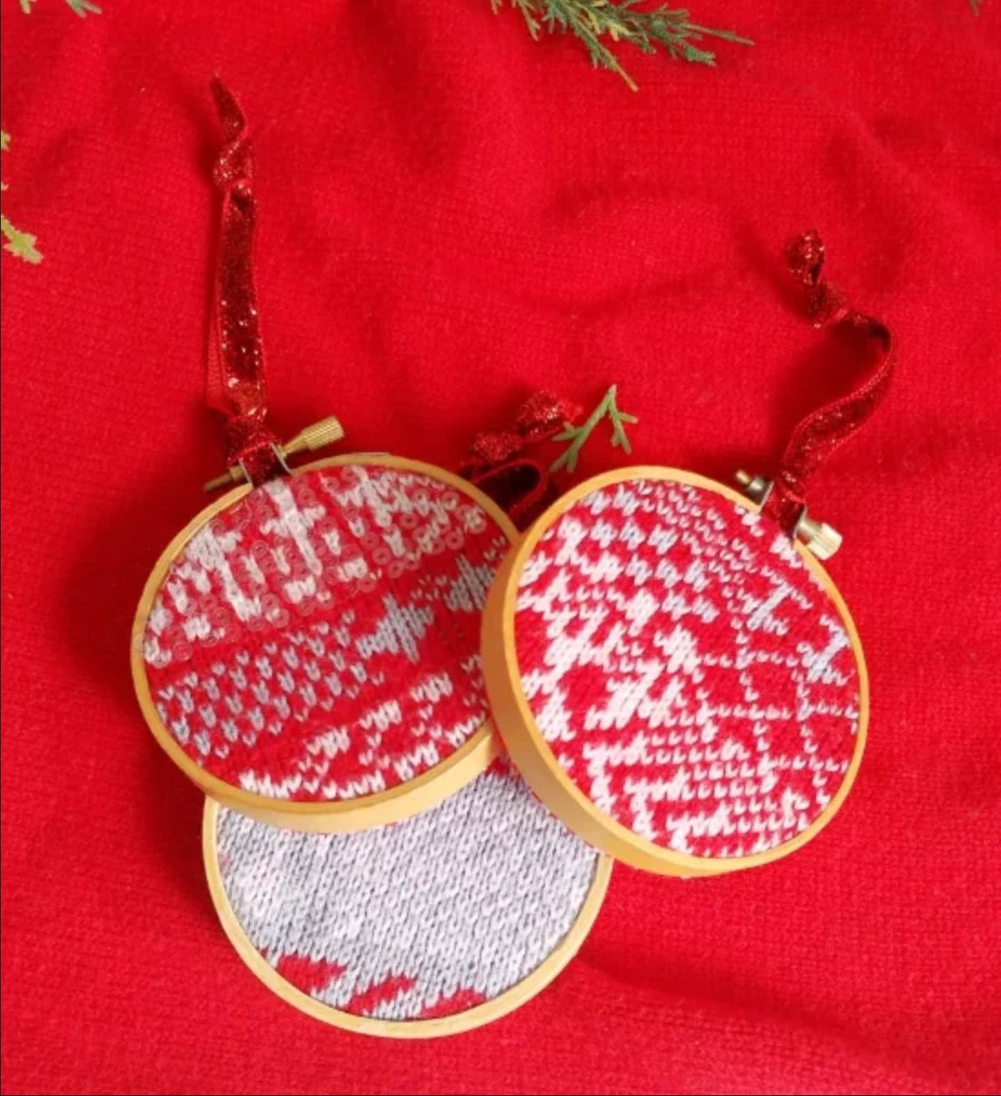 Three small embroidery hoops with red, white, and gray knitted patterns and red ribbon hangers are displayed on a red fabric background with greenery at the top.