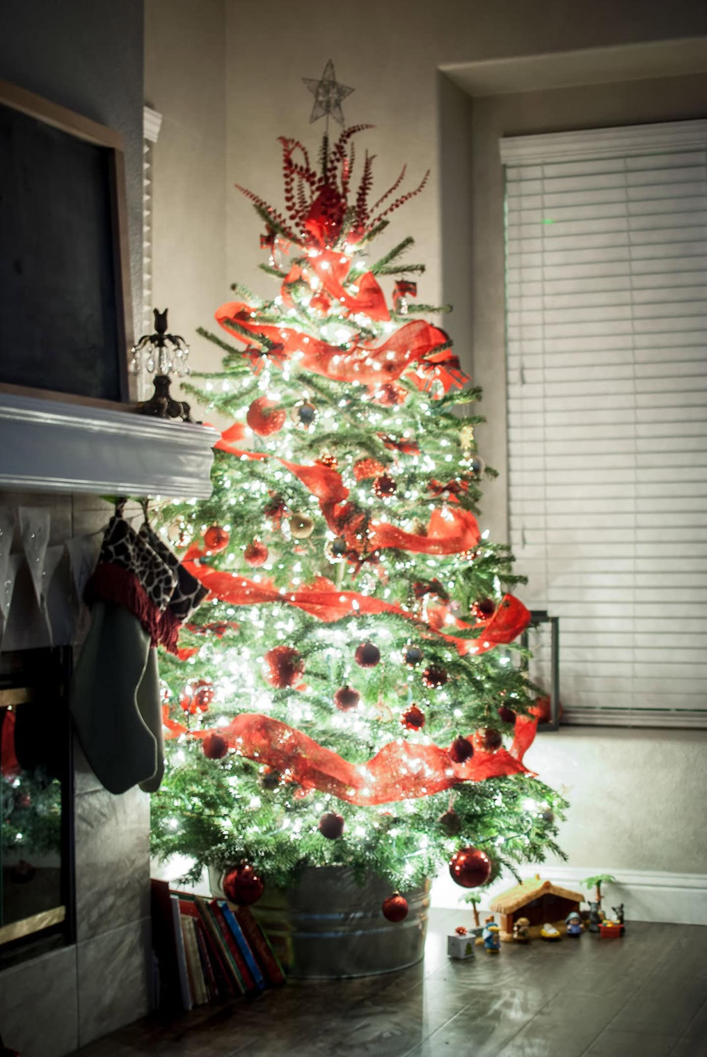 A decorated Christmas tree with red ribbons and ornaments stands by a window next to a fireplace and a small Nativity scene at the base.