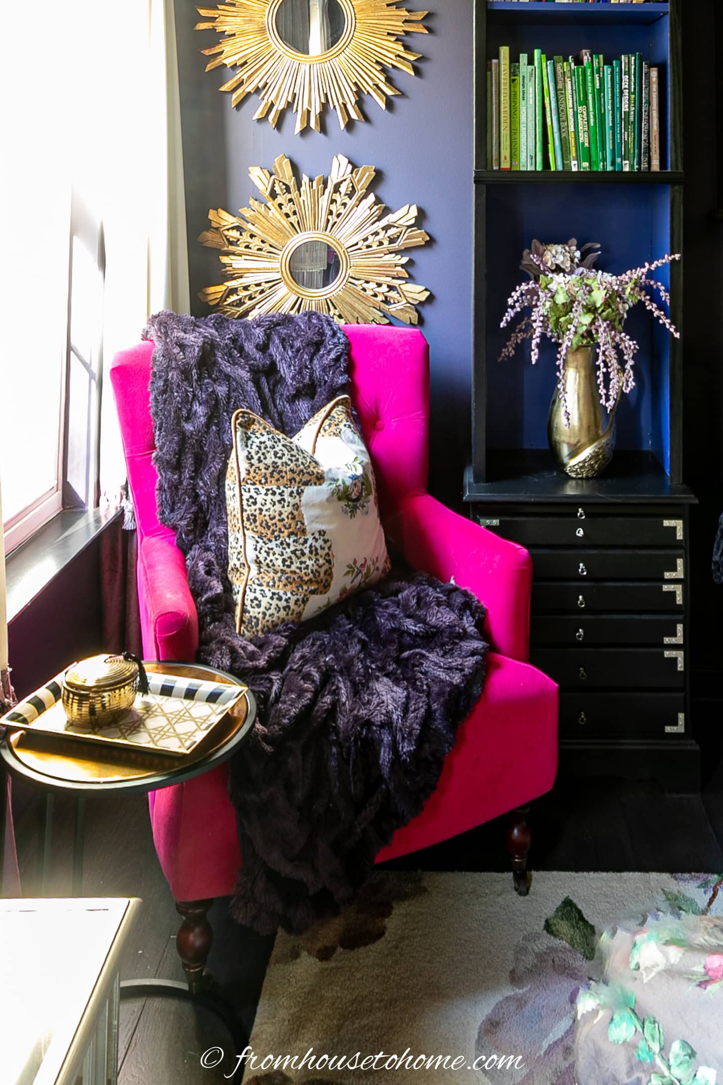 A bright pink armchair with a cheetah print pillow and dark throw sits beside a small table and black shelving with green books and a floral vase. Sunlight streams through a nearby window.