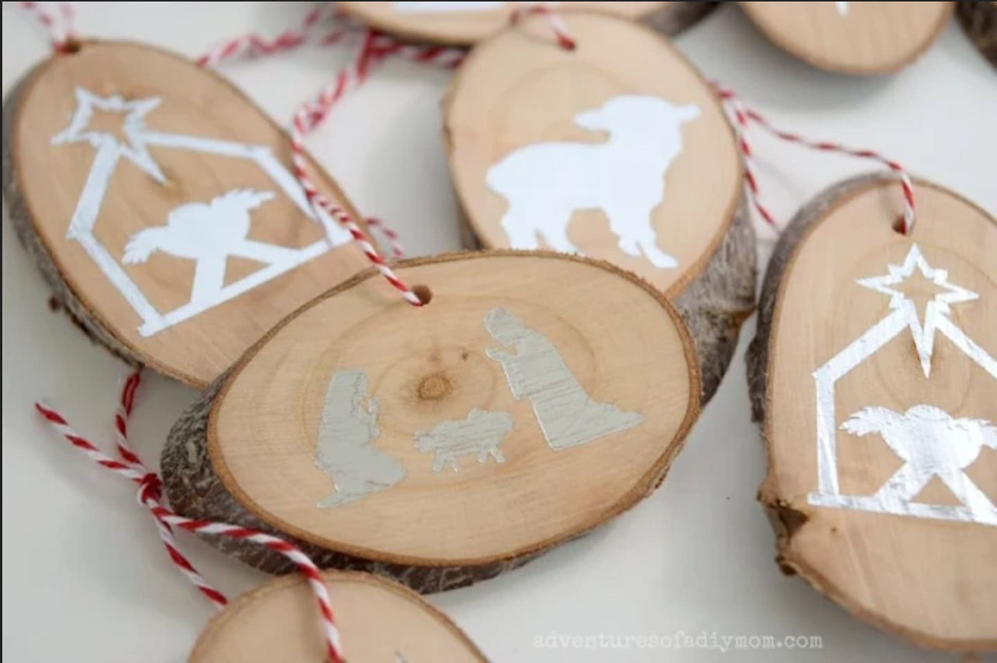 Wood slice ornaments with silver nativity scene and lamb silhouettes, each hung with red and white twine.