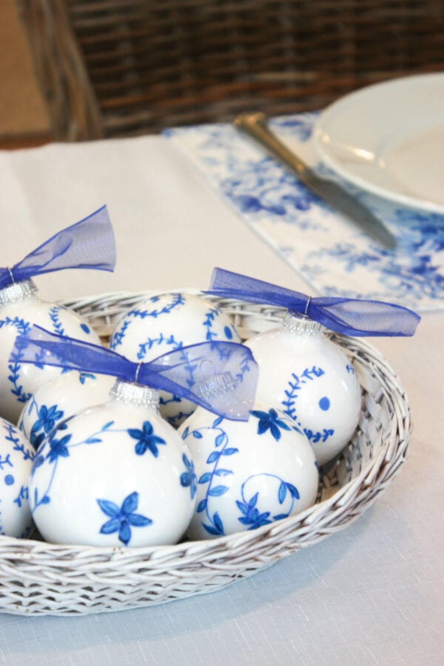 A white basket filled with white ornaments decorated with blue floral patterns and topped with purple ribbons, placed on a white tablecloth near a plate and cutlery.