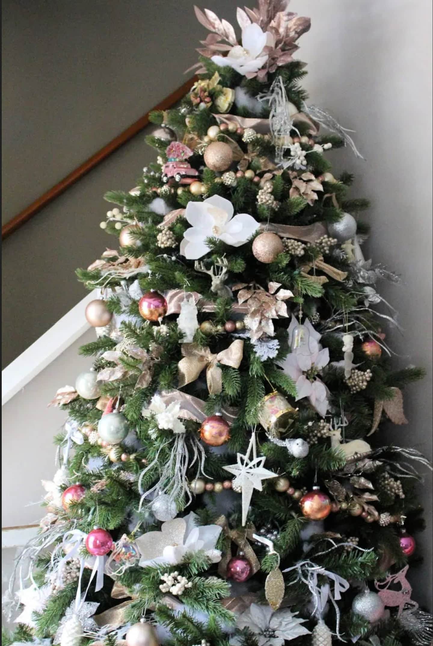 A decorated Christmas tree with pink, gold, and white ornaments, artificial flowers, and a large floral tree topper stands near a staircase.