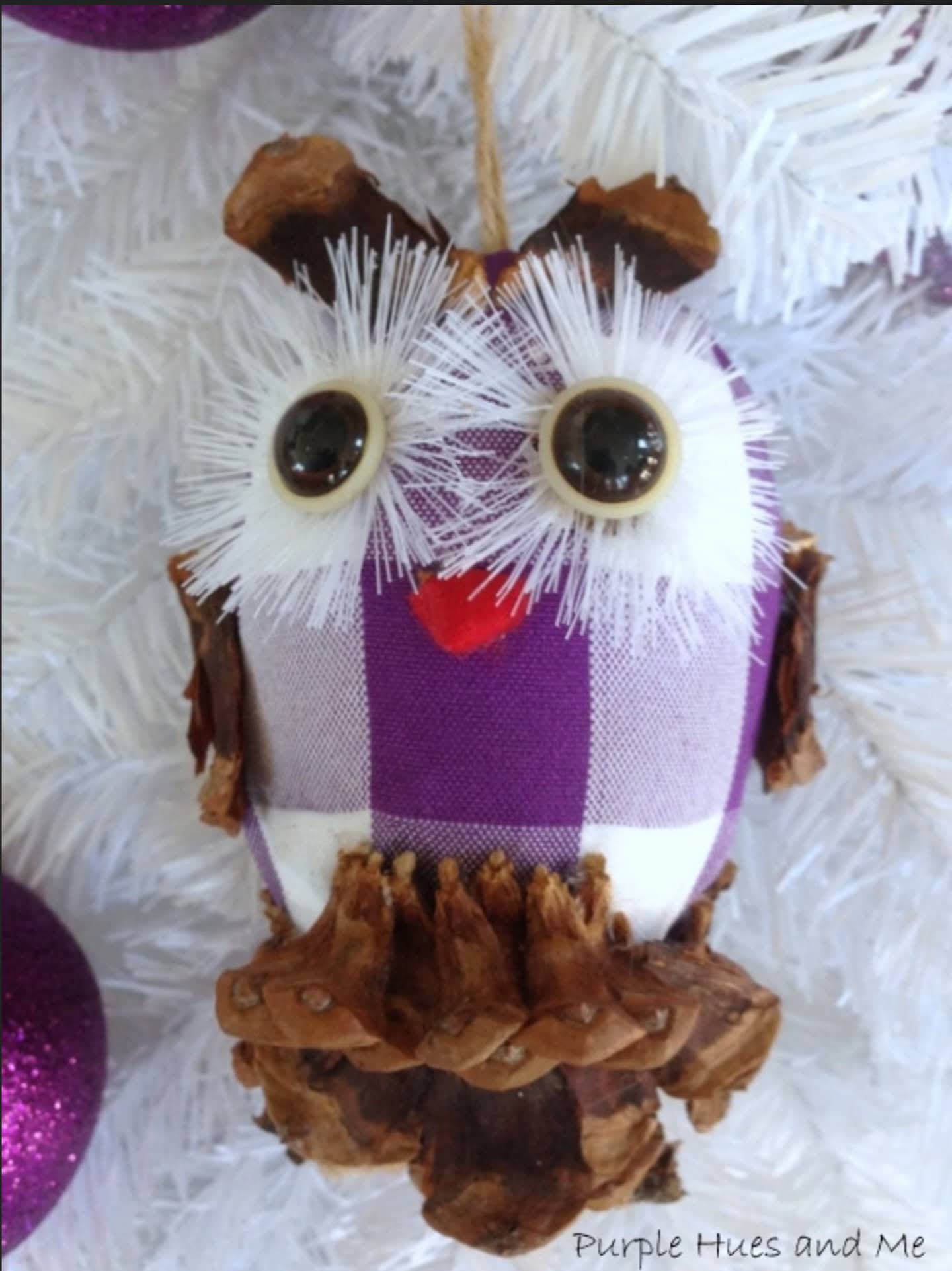 Handmade owl ornament made from pinecones, a purple and white checkered fabric, plastic eyes, and white bristles, hanging on a white Christmas tree.
