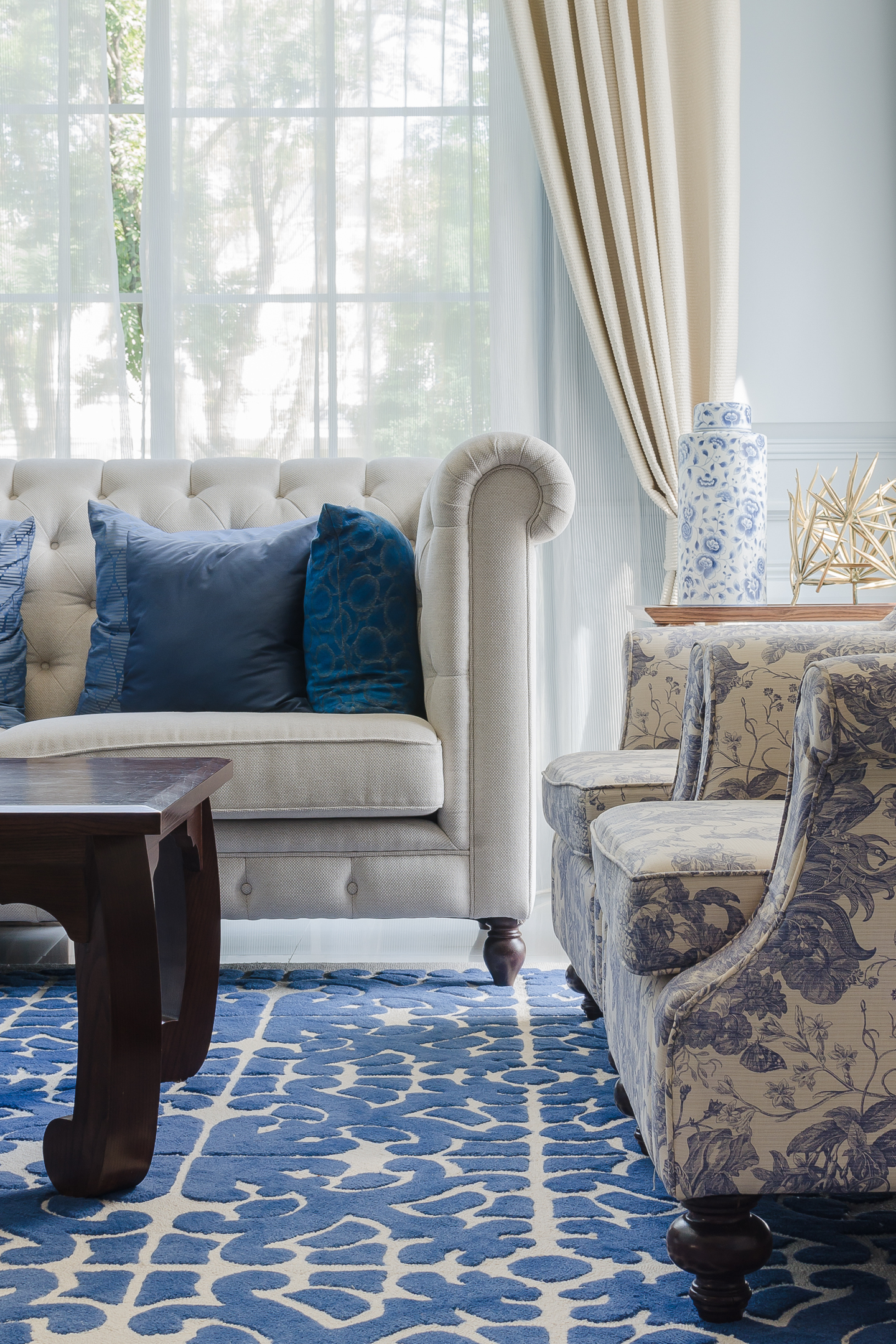 A living room with a beige tufted sofa, blue accent pillows, patterned armchairs, a dark wood coffee table, a blue and white rug, and sunlight filtering through sheer curtains.