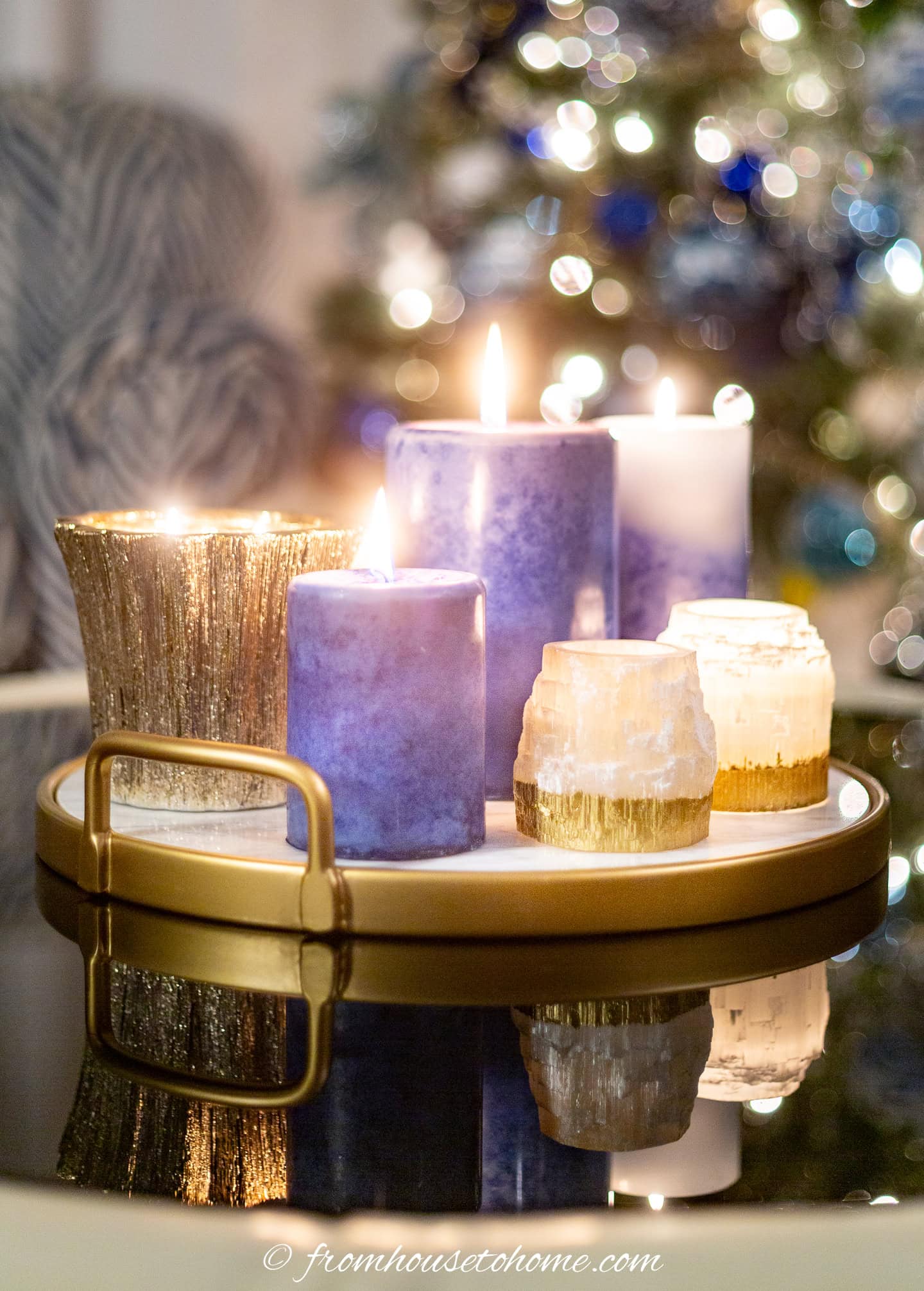 A gold tray displays lit purple candles and white votives with gold bases on a reflective table, with a decorated Christmas tree blurred in the background.