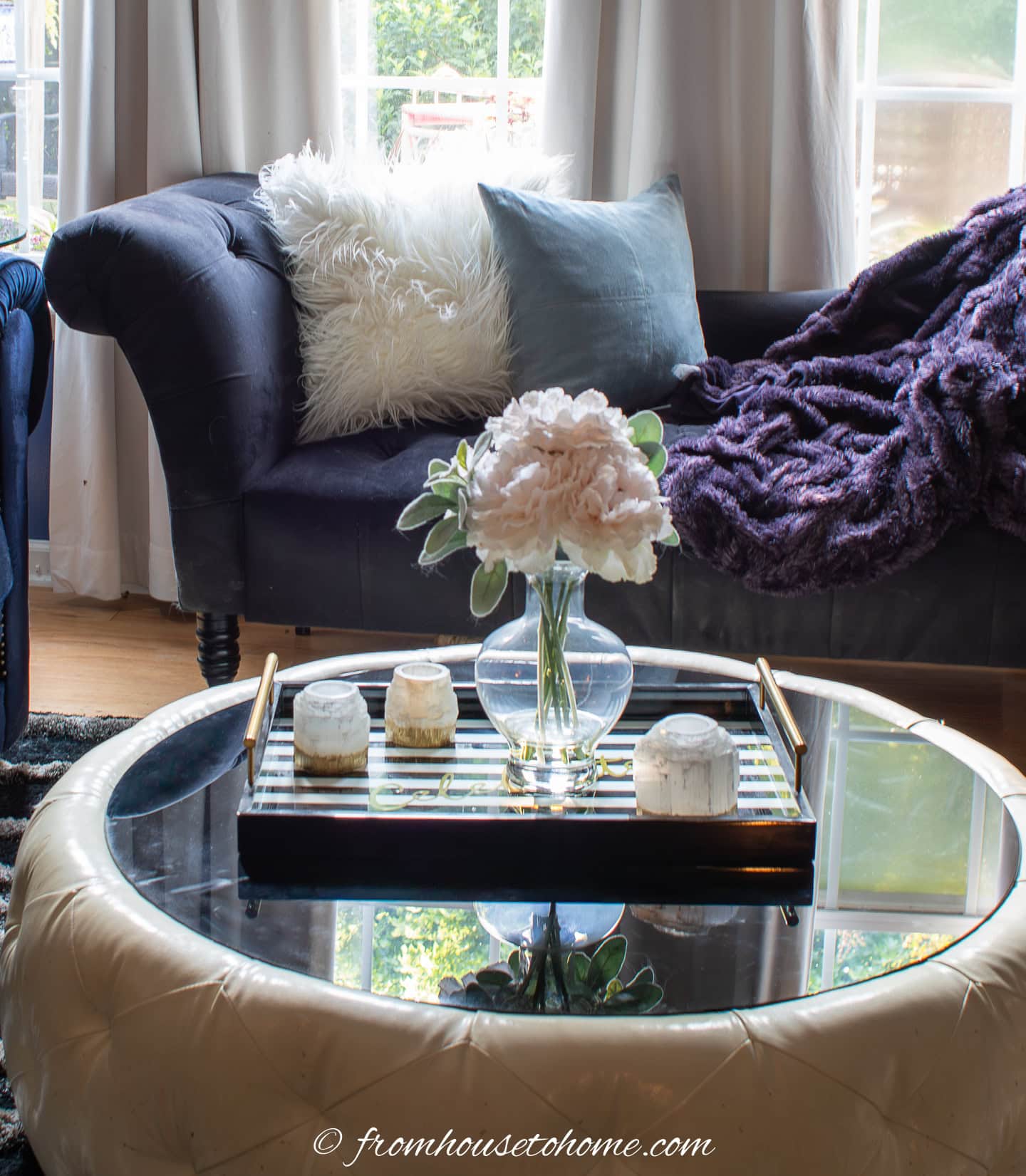 A glass coffee table with a vase of light pink flowers and decorative objects on a tray, in front of a dark sofa with white and purple pillows and a blanket.