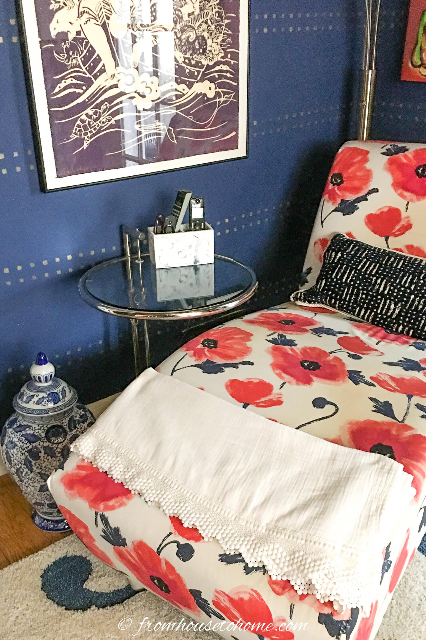 A chaise lounge with red and blue floral upholstery next to a glass side table, a black and white pillow, a white throw blanket, and a blue-and-white ceramic jar.