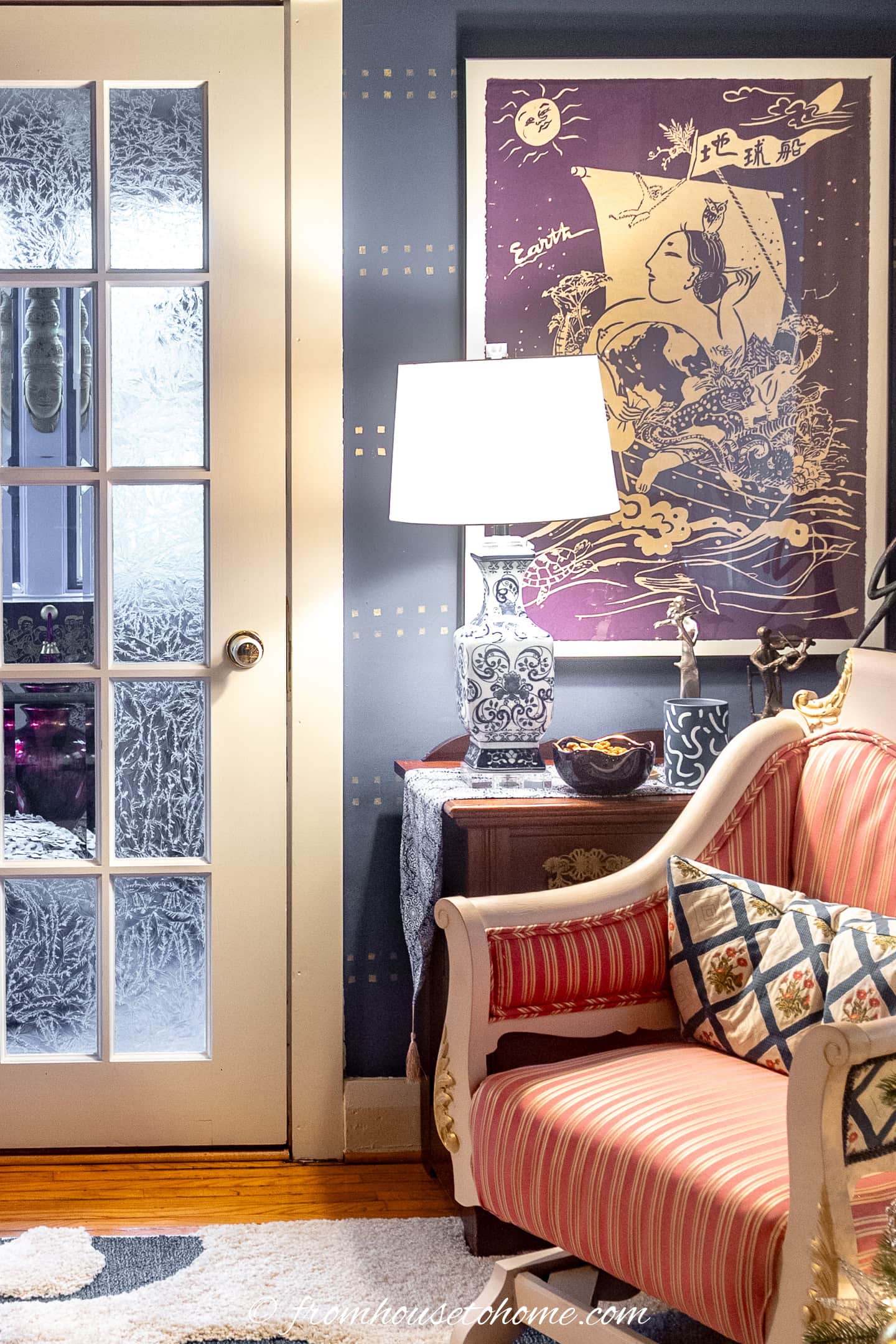 A cozy living room corner with a striped armchair, patterned pillows, side table with lamp, decorative objects, a large Asian-inspired wall art, and a glass door with frosted designs.