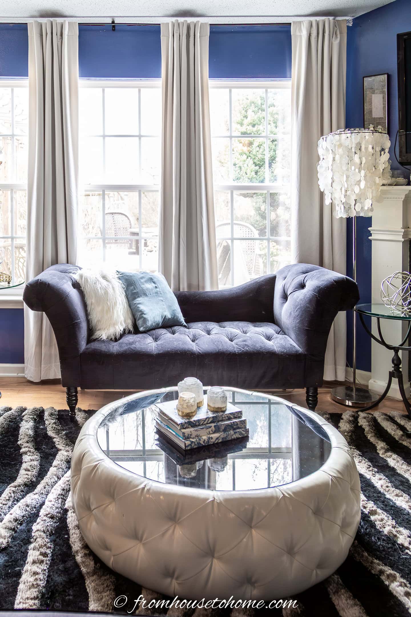 A dark blue tufted chaise sits in front of large windows with floor-length curtains; a round, tufted ottoman with a glass top serves as a coffee table.