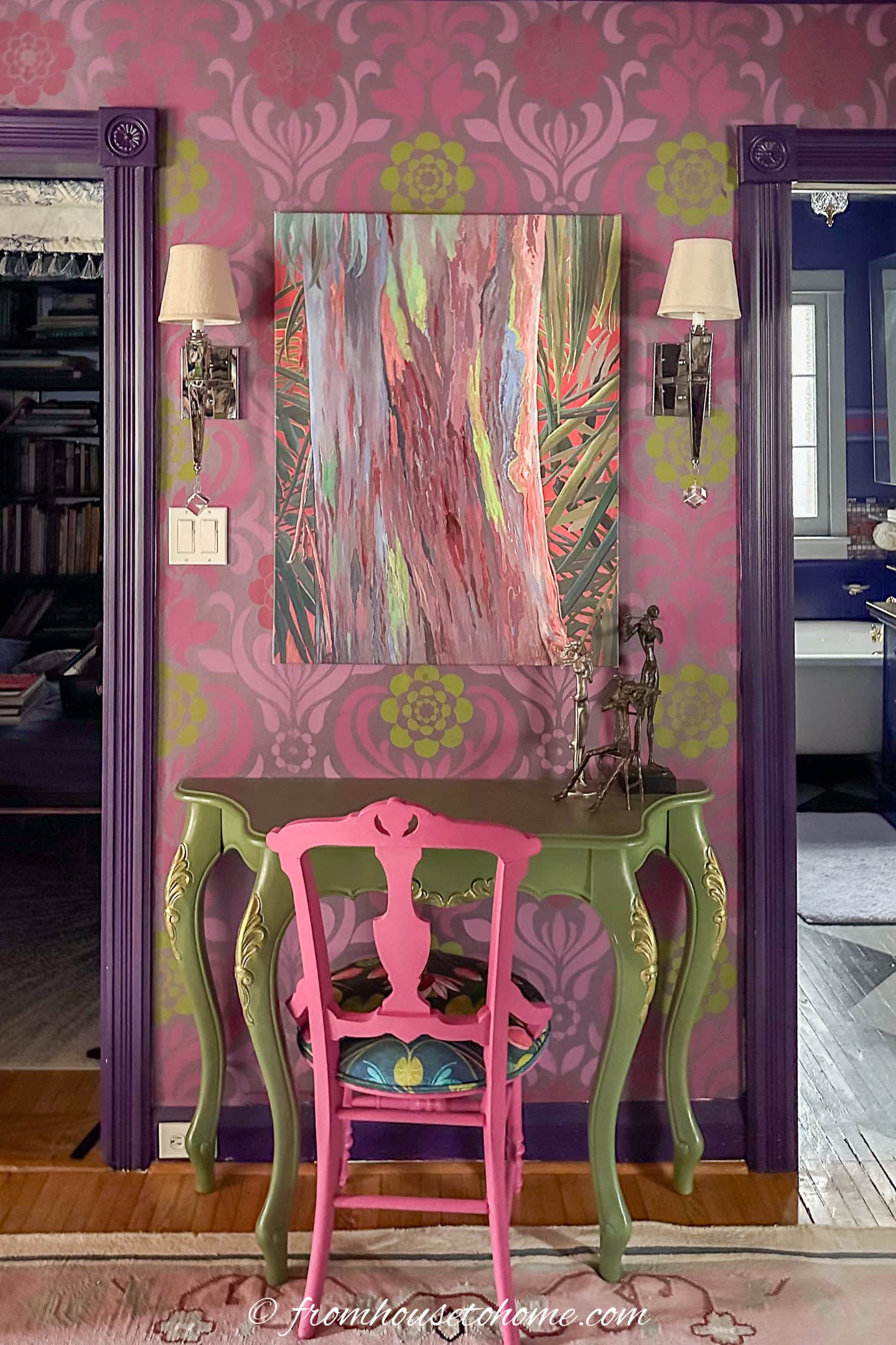 A small decorative desk with a pink chair sits against a purple and pink patterned wall, beneath an abstract painting and two wall sconces.