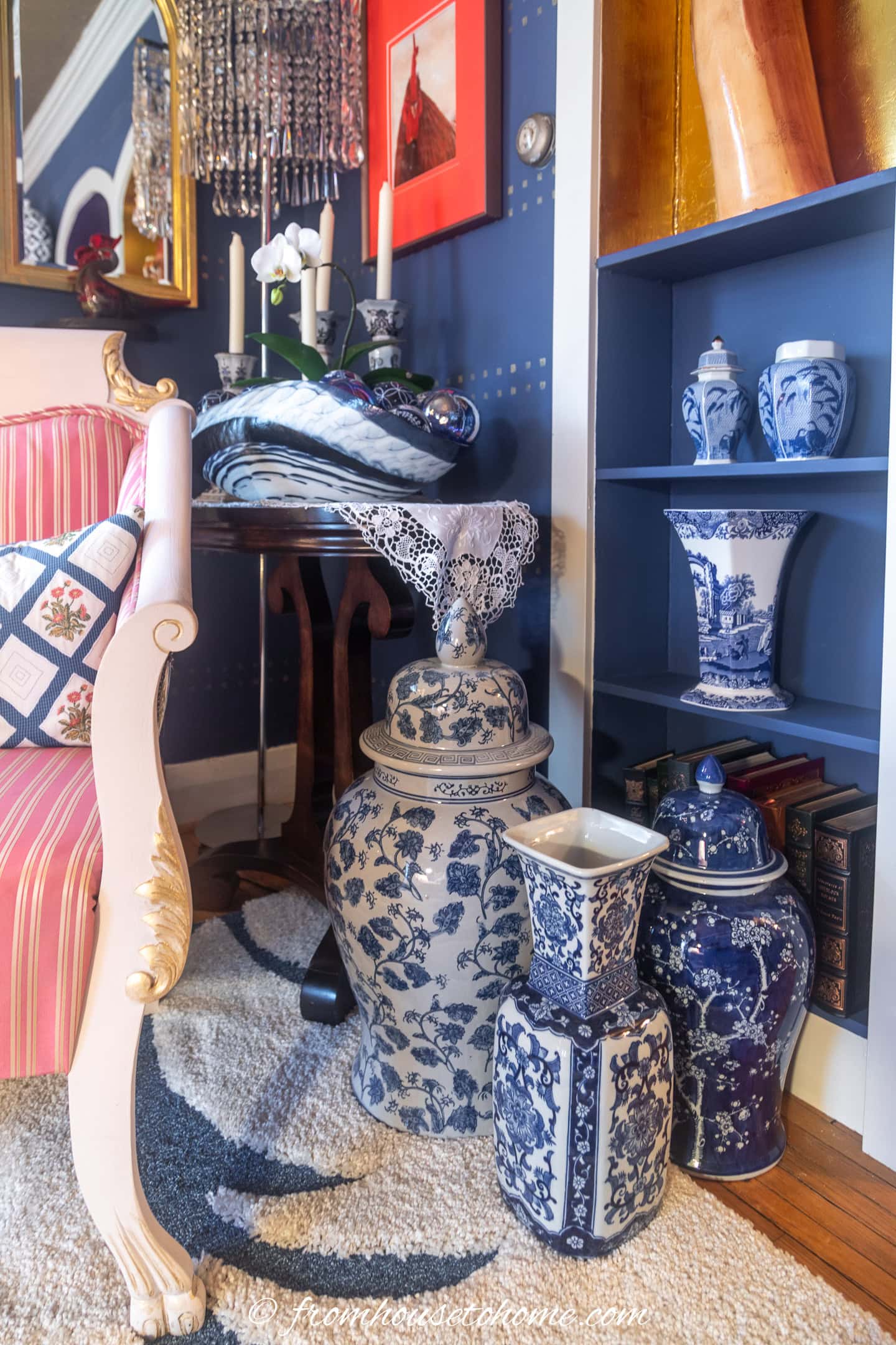 A collection of blue and white porcelain vases and jars arranged next to a pink striped chair and a blue bookshelf in a decorated living room corner.