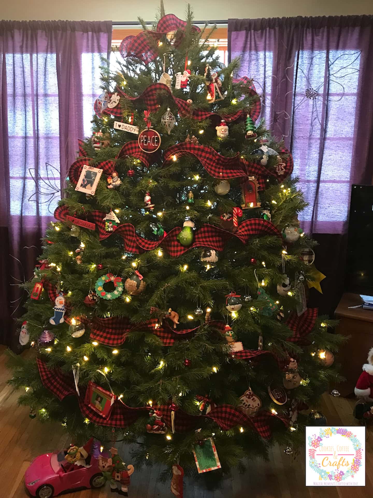 A decorated Christmas tree with red plaid ribbon, various ornaments, and string lights stands in front of purple curtains.
