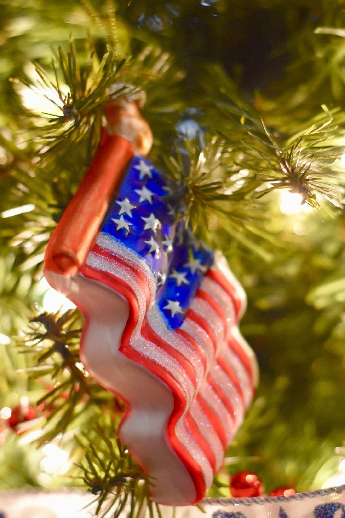 Close-up of an American flag ornament hanging on a Christmas tree, surrounded by green pine needles and lit by soft white lights.