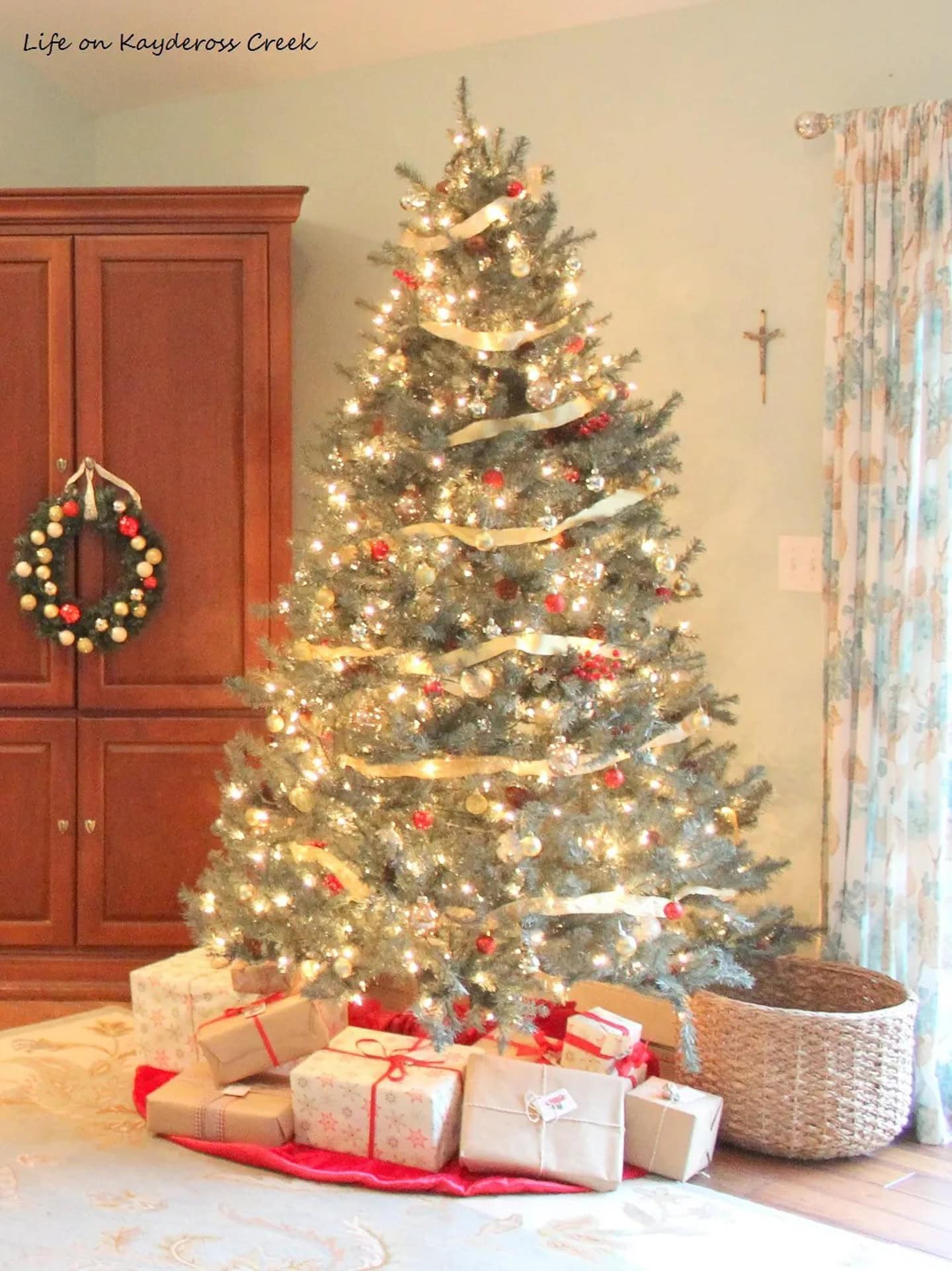A decorated Christmas tree with white lights, gold ribbon, red and gold ornaments, and wrapped gifts underneath stands in a living room near a basket and curtained window.