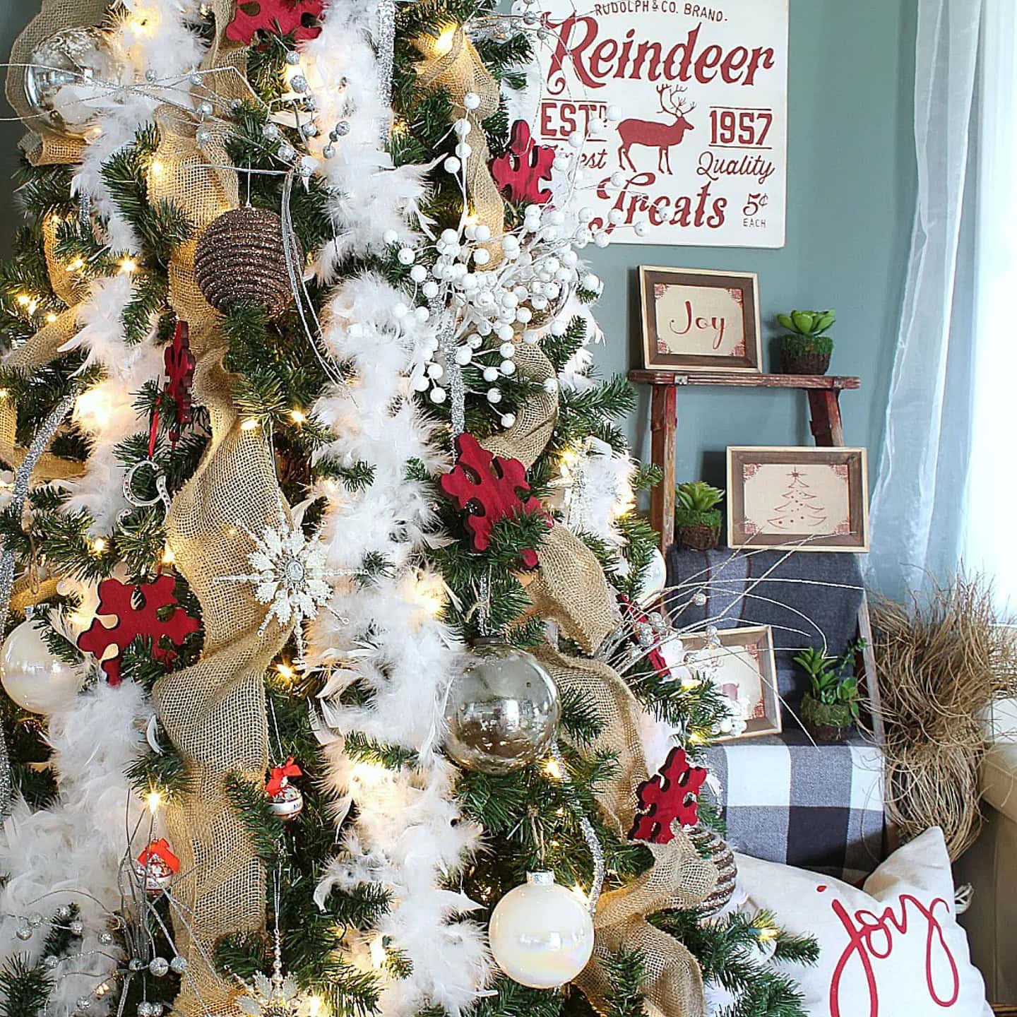 A Christmas tree decorated with burlap ribbon, white feathers, red ornaments, and glass balls stands next to festive framed signs and a