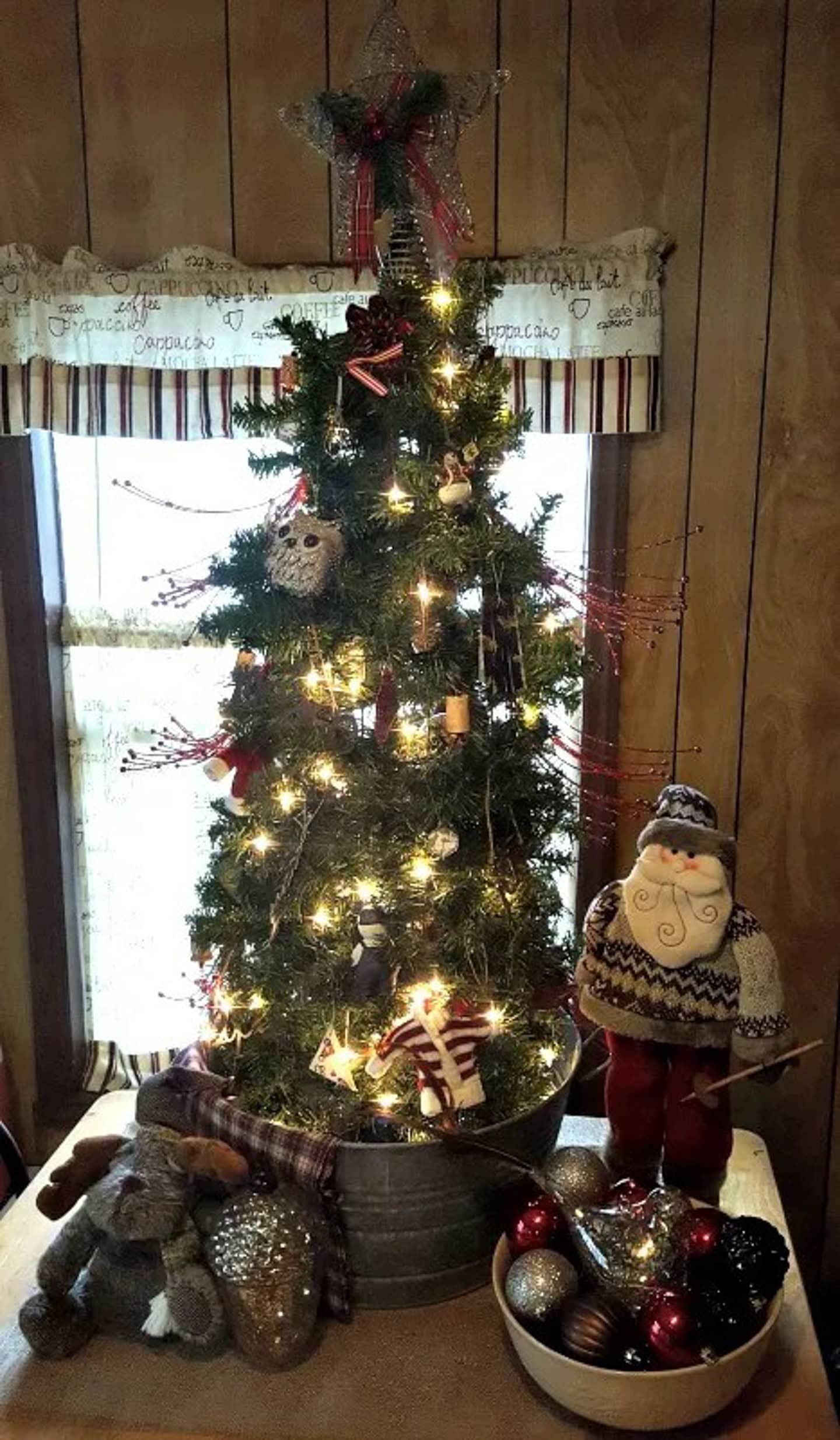 A small decorated Christmas tree with lights stands in a metal tub on a table. Beside it are a Santa figurine, a stuffed toy, and a bowl of assorted ornaments.