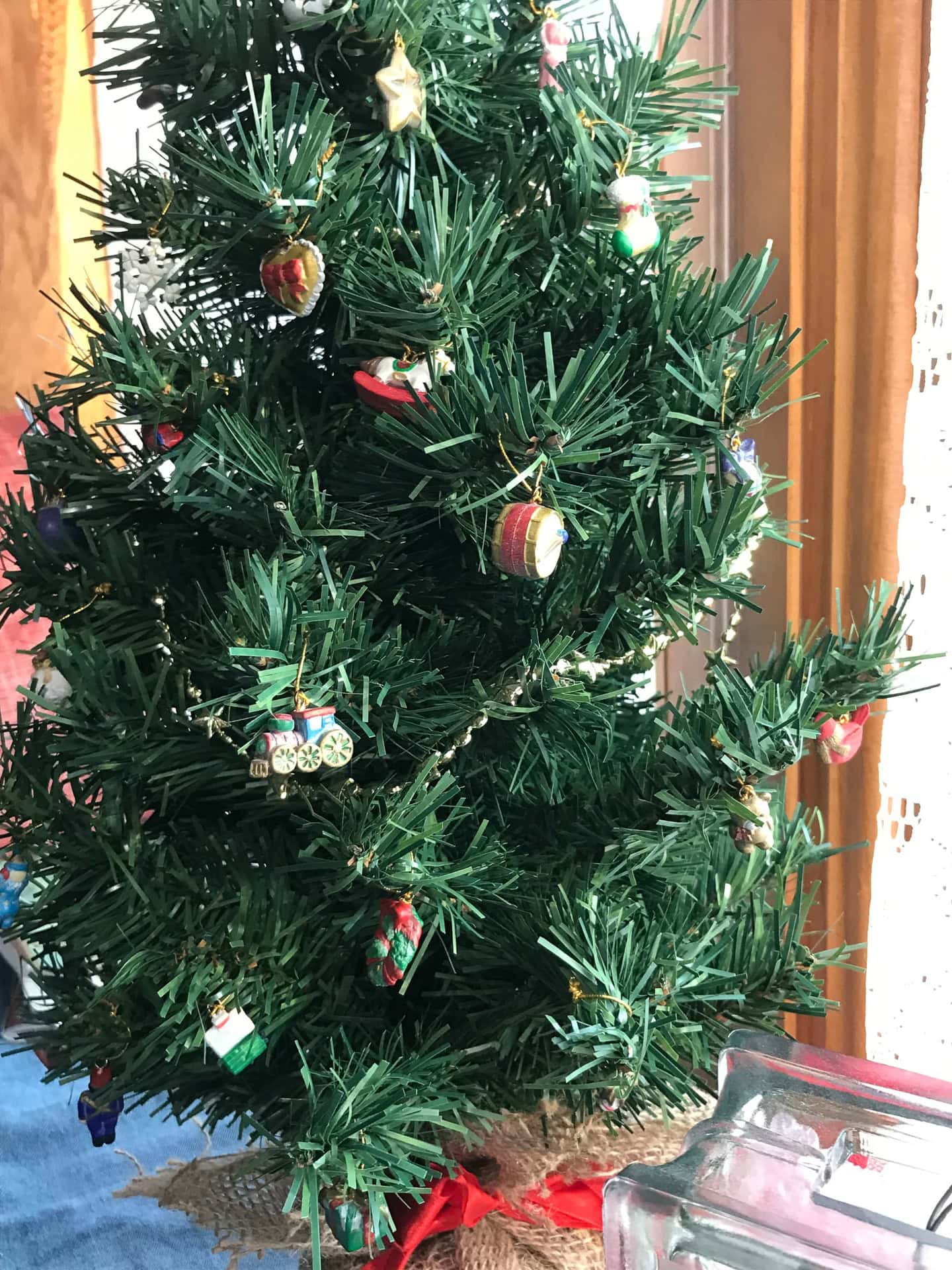 A small artificial Christmas tree decorated with miniature ornaments stands on a table near a window with lace curtains.