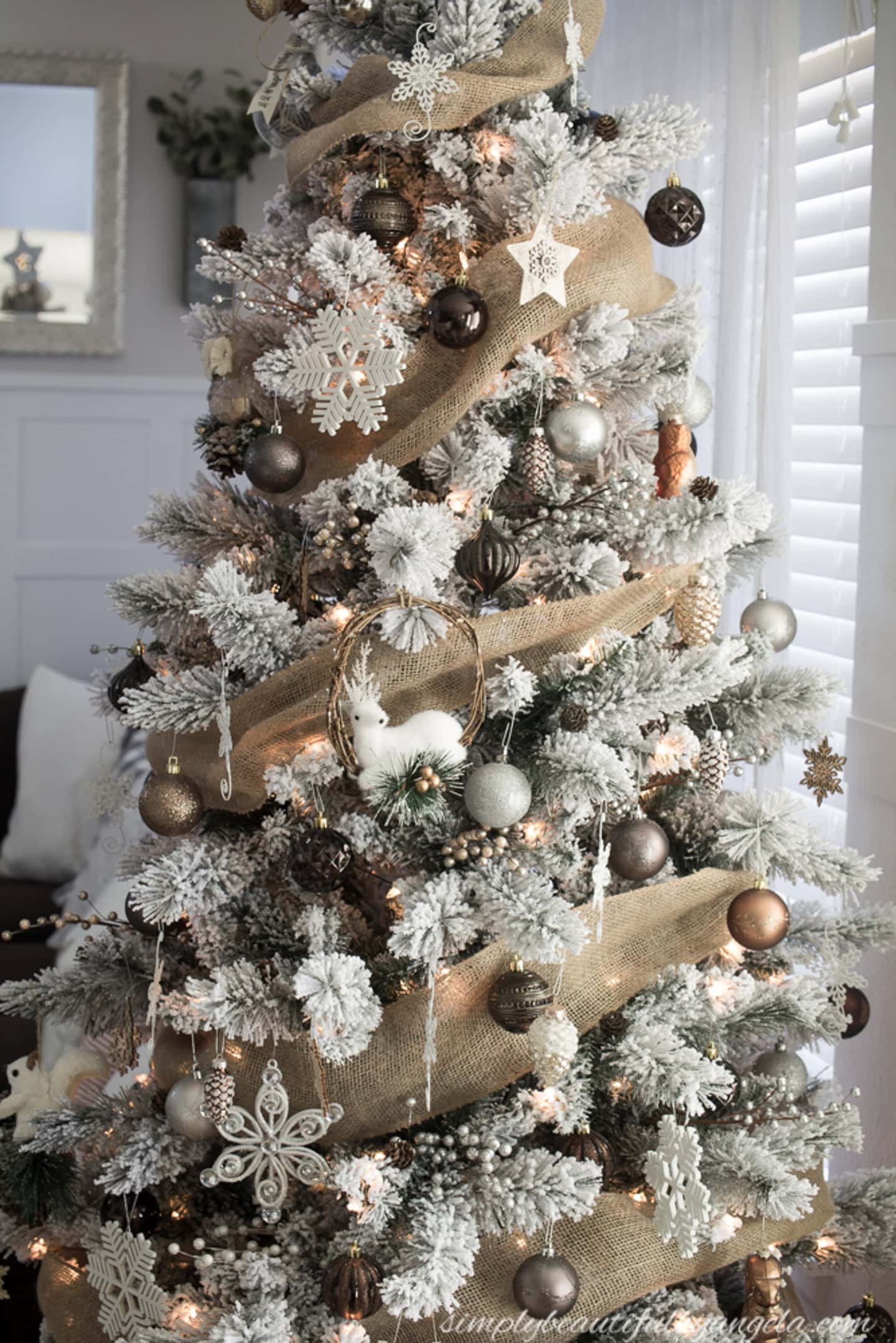 A flocked Christmas tree decorated with burlap ribbon, white and brown ornaments, snowflakes, and warm white lights stands near a window.