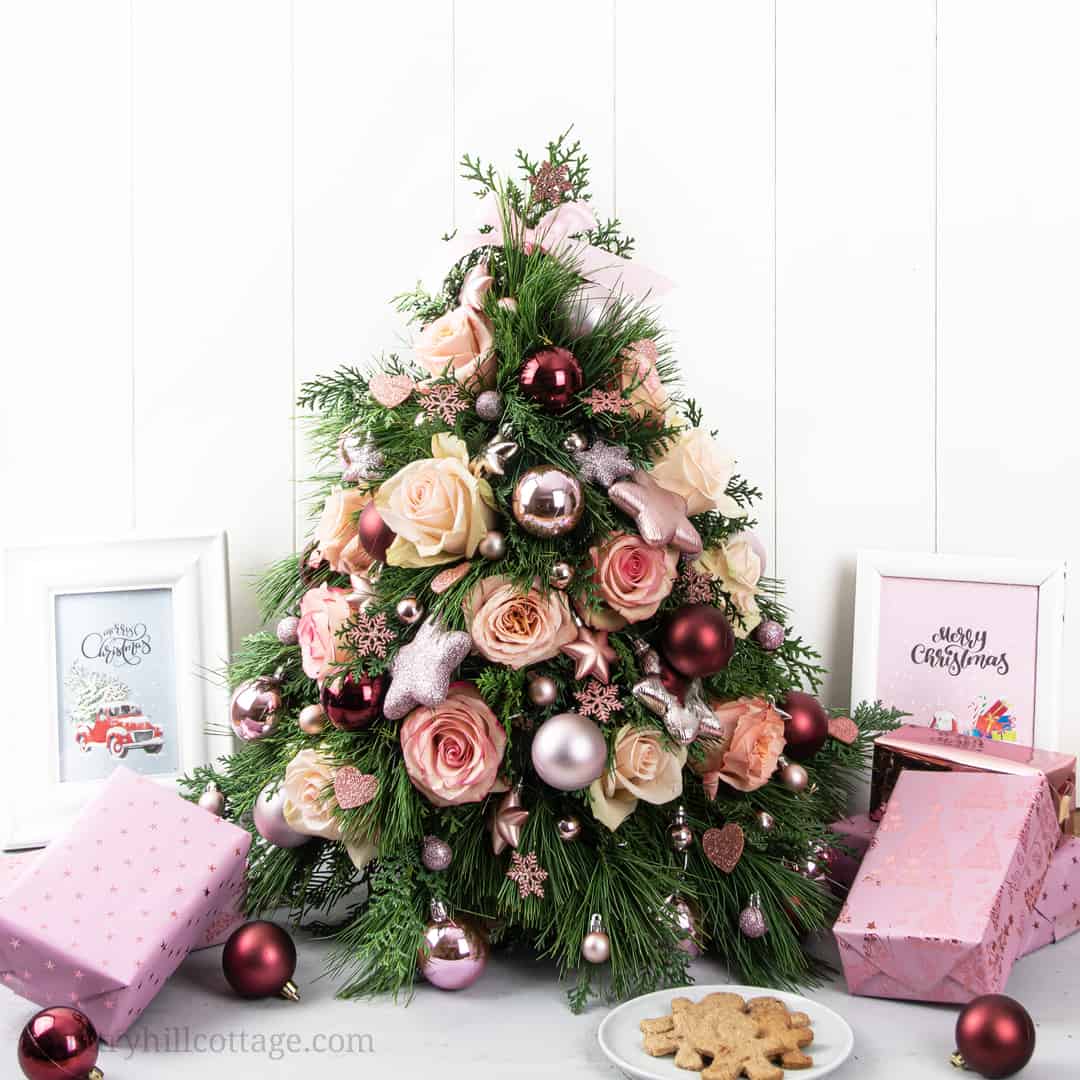 A small artificial Christmas tree decorated with pink roses, ornaments, and surrounded by framed prints, wrapped gifts, and a plate of cookies on a white background.