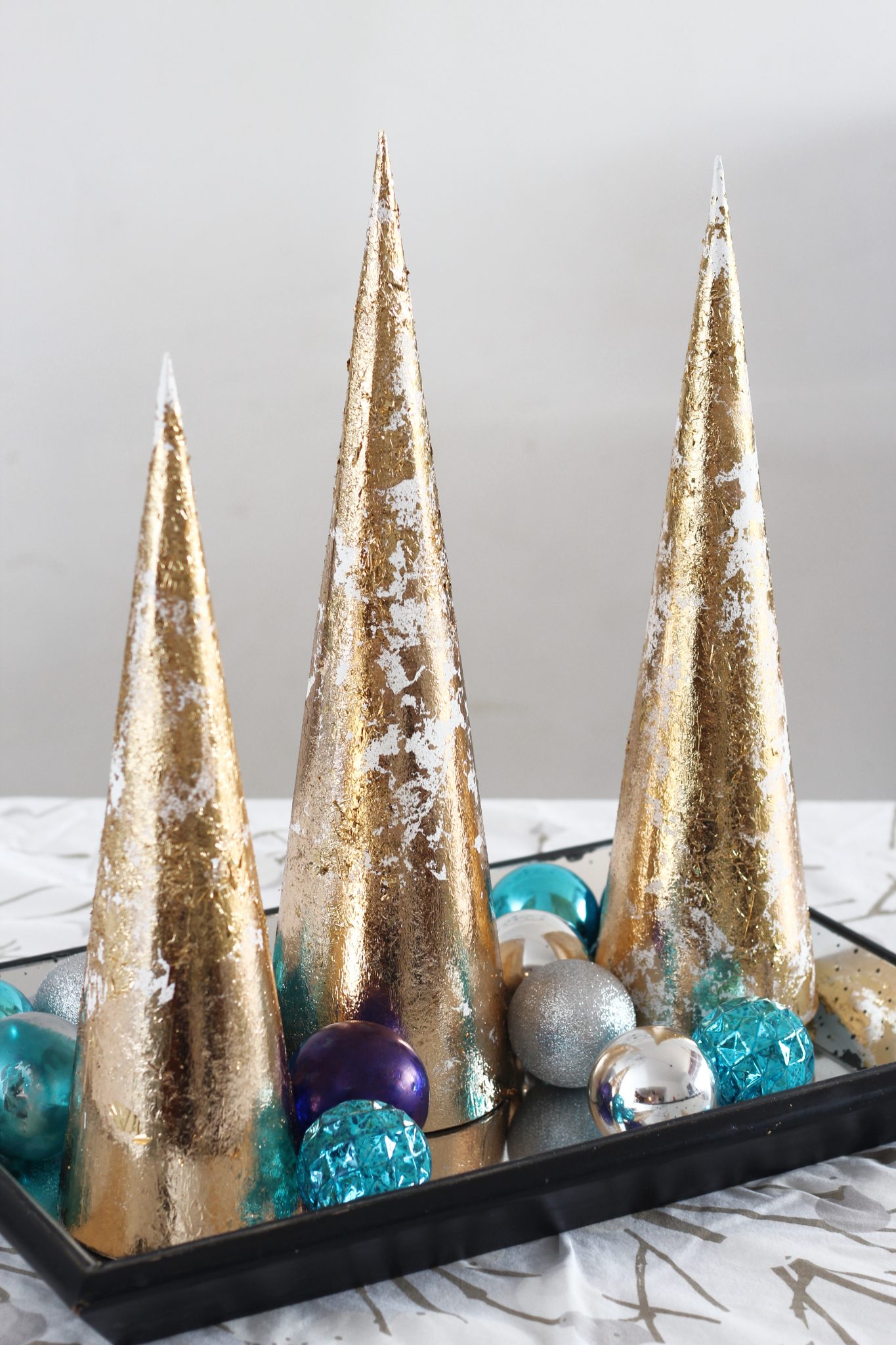 Three gold cone-shaped decorations sit on a black tray, surrounded by blue, silver, and purple ornaments on a white patterned surface.