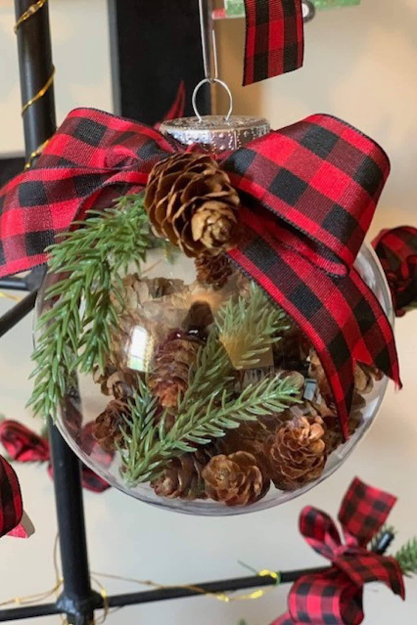 A clear Christmas ornament filled with pinecones and evergreen sprigs, topped with a red and black plaid bow, hanging from a black metal stand.