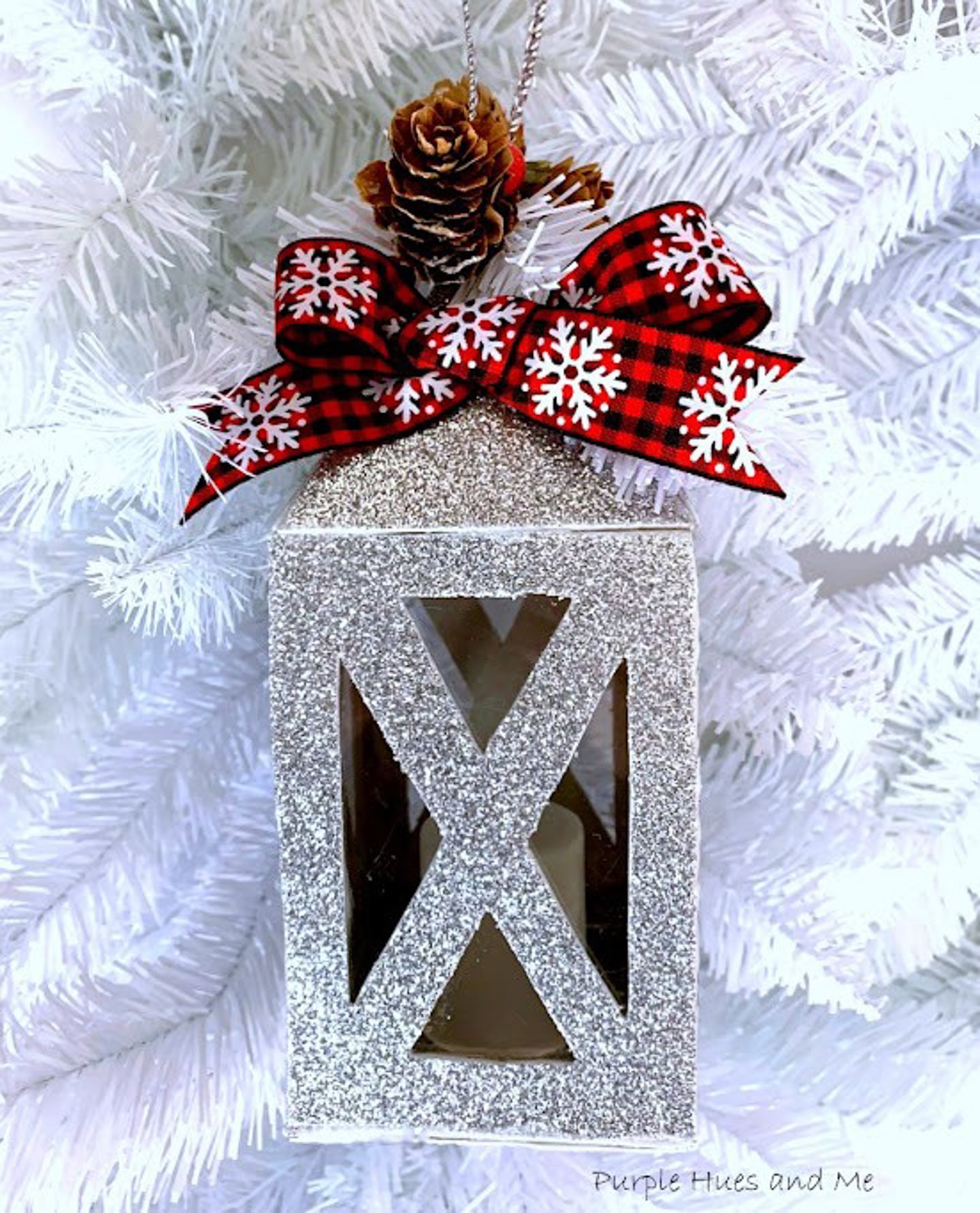 A silver glitter lantern ornament hangs on a white Christmas tree, topped with a pine cone, red plaid bow, and snowflake decorations.
