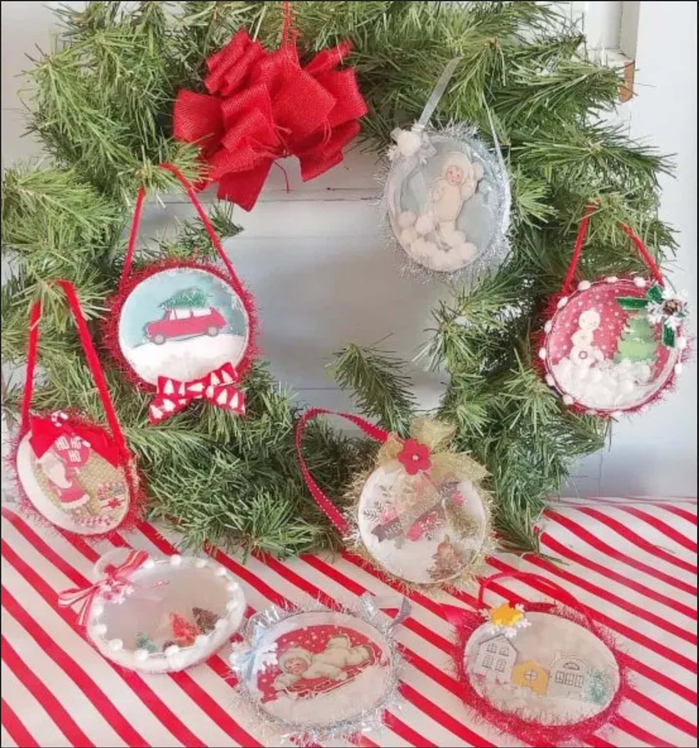 A green wreath with a red bow is decorated with handmade round ornaments featuring various holiday scenes, displayed on a red and white striped surface.
