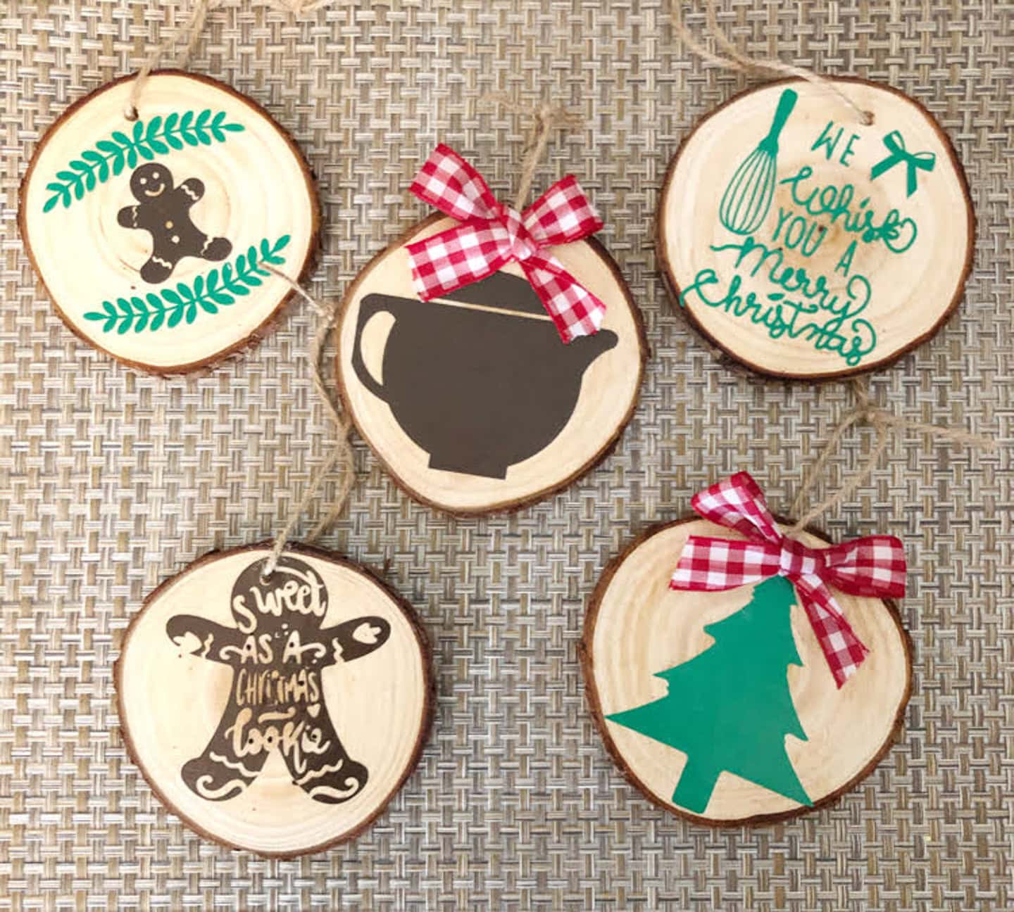 Five round wooden ornaments with holiday-themed designs and red plaid bows displayed on a woven surface. Designs include a gingerbread man, tree, bowl, whisk, and festive text.