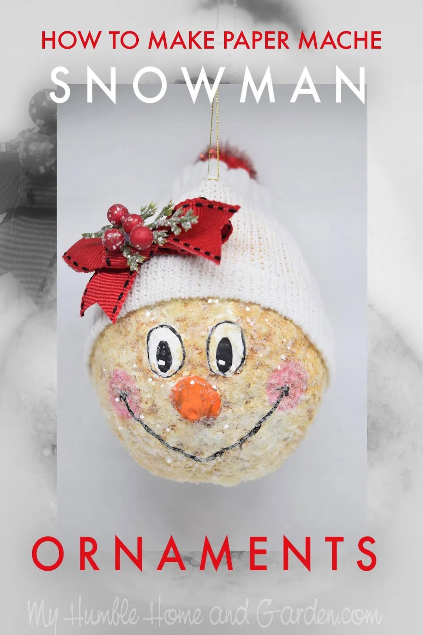 A paper mache snowman ornament with a red bow, white hat, painted face, and orange nose hangs against a white background. Text reads
