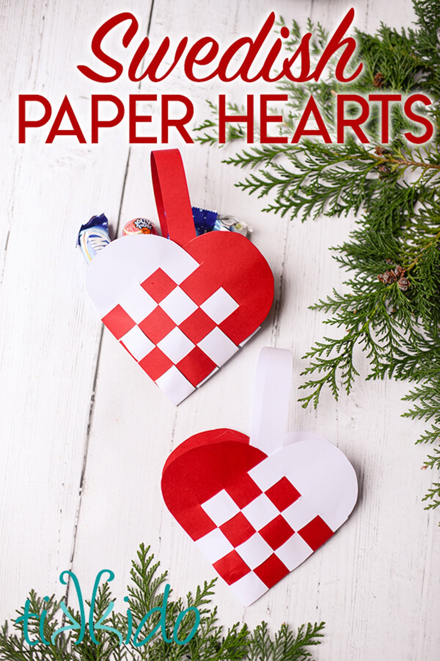 Two woven red and white paper heart decorations with handles, filled with candy, are displayed on a white wooden surface with pine branches nearby. Text reads