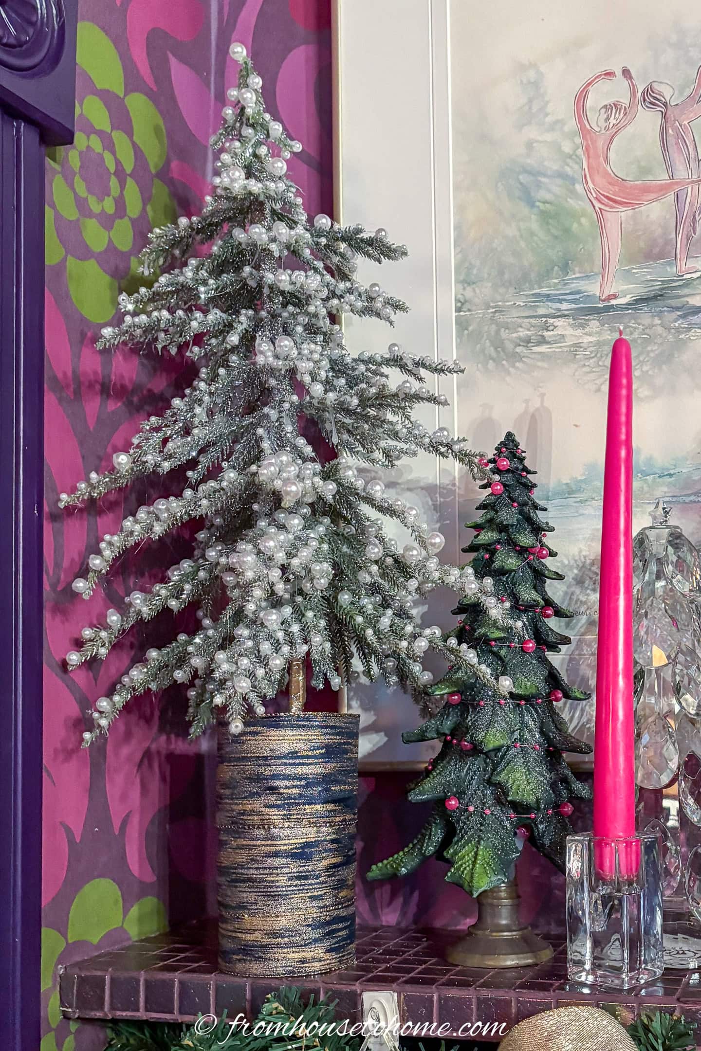 Two decorative artificial Christmas trees, one tall and snowy in a blue pot, and one smaller with red ornaments, stand beside a pink taper candle on a patterned surface.