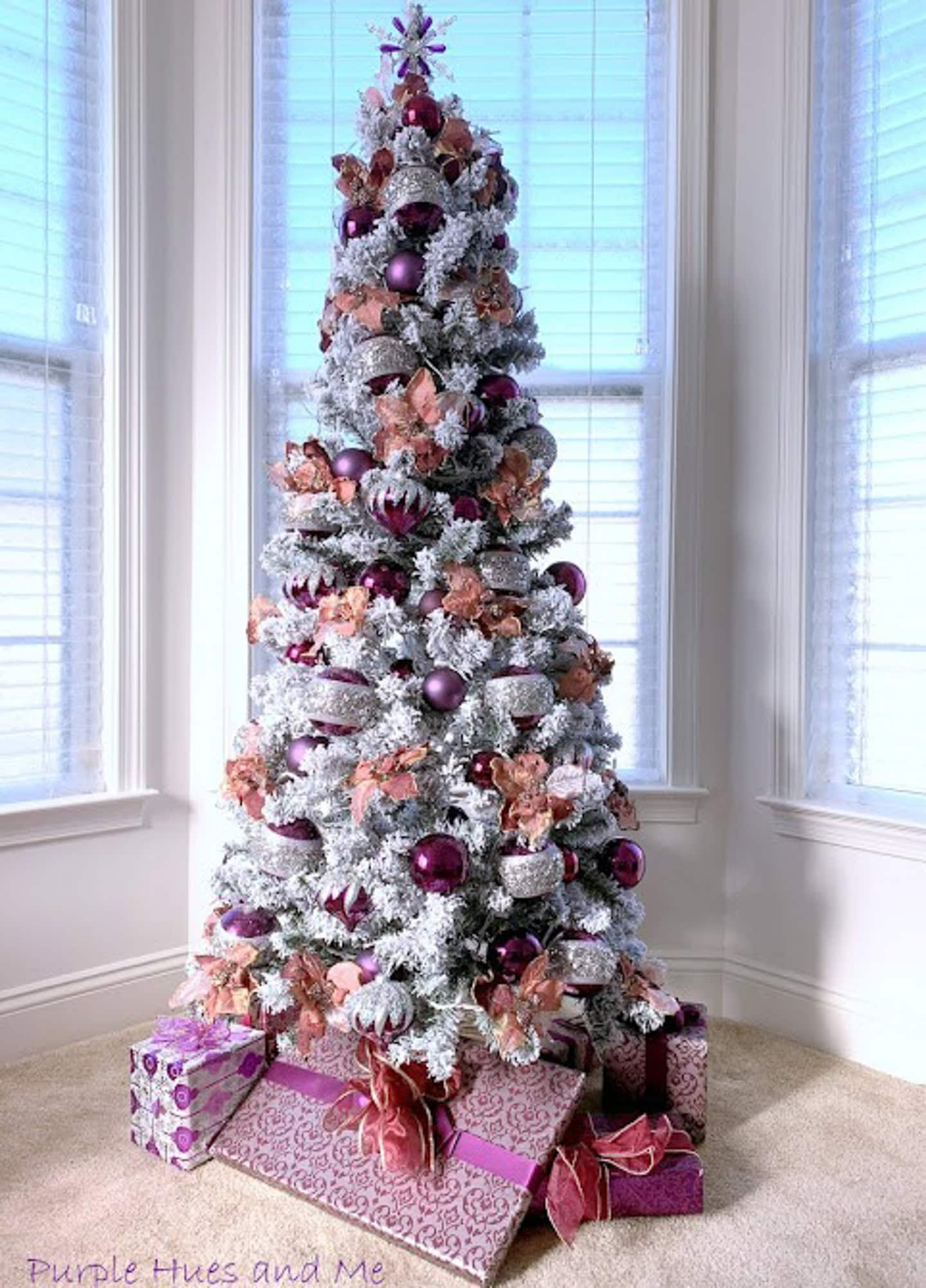 A flocked Christmas tree decorated with purple and pink ornaments stands in front of windows, with wrapped gifts in matching colors arranged at its base.
