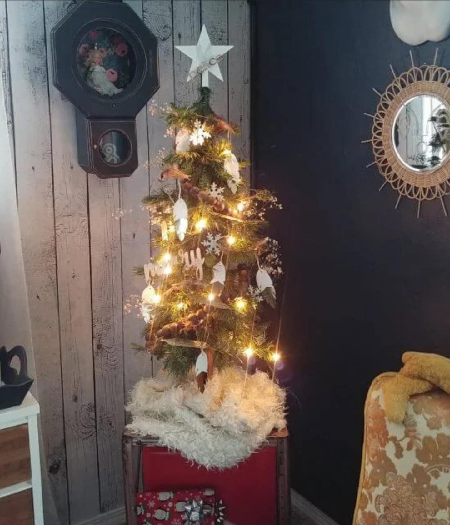 A small decorated Christmas tree with lights and white ornaments stands on a red box, next to a vintage chair and wall decor in a cozy room.