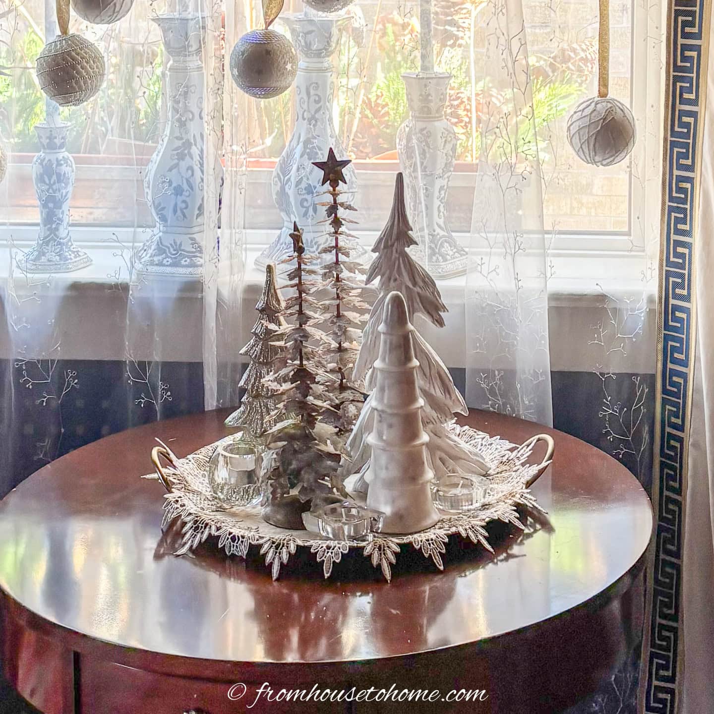 a group of mini christmas trees on a table in front of a window decorated with white ornaments hung from gold ribbon