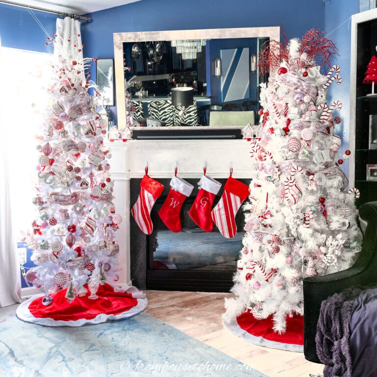 Two white Christmas trees decorated with red and white ornaments stand on either side of a fireplace with three red stockings hanging from the mantel in a blue-walled room.