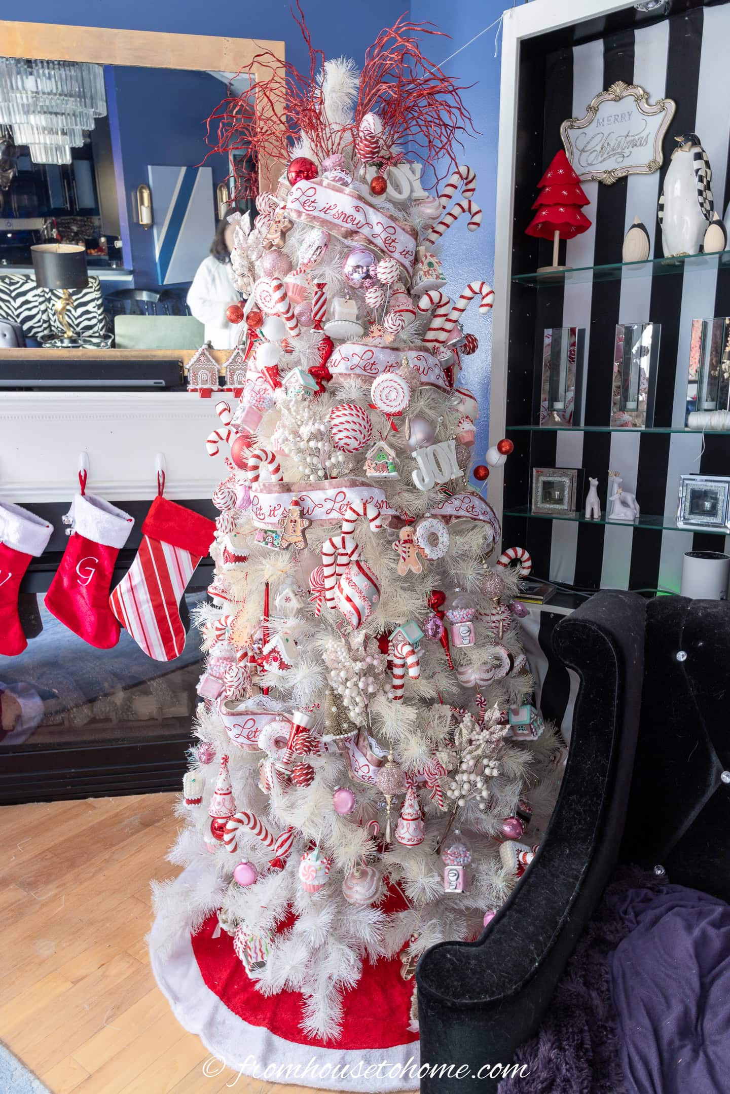 A white Christmas tree decorated with red and white ornaments, peppermint-themed decorations, and a red tree skirt, stands beside a fireplace with hanging stockings.