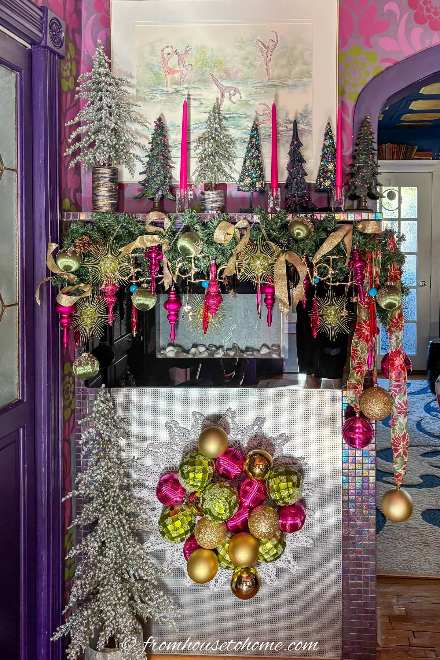 An electric fireplace decorated with mini Christmas trees, candles, a garland and a Christmas ornament wall hanging all decorated with fuchsia, lime green and gold Christmas home decor