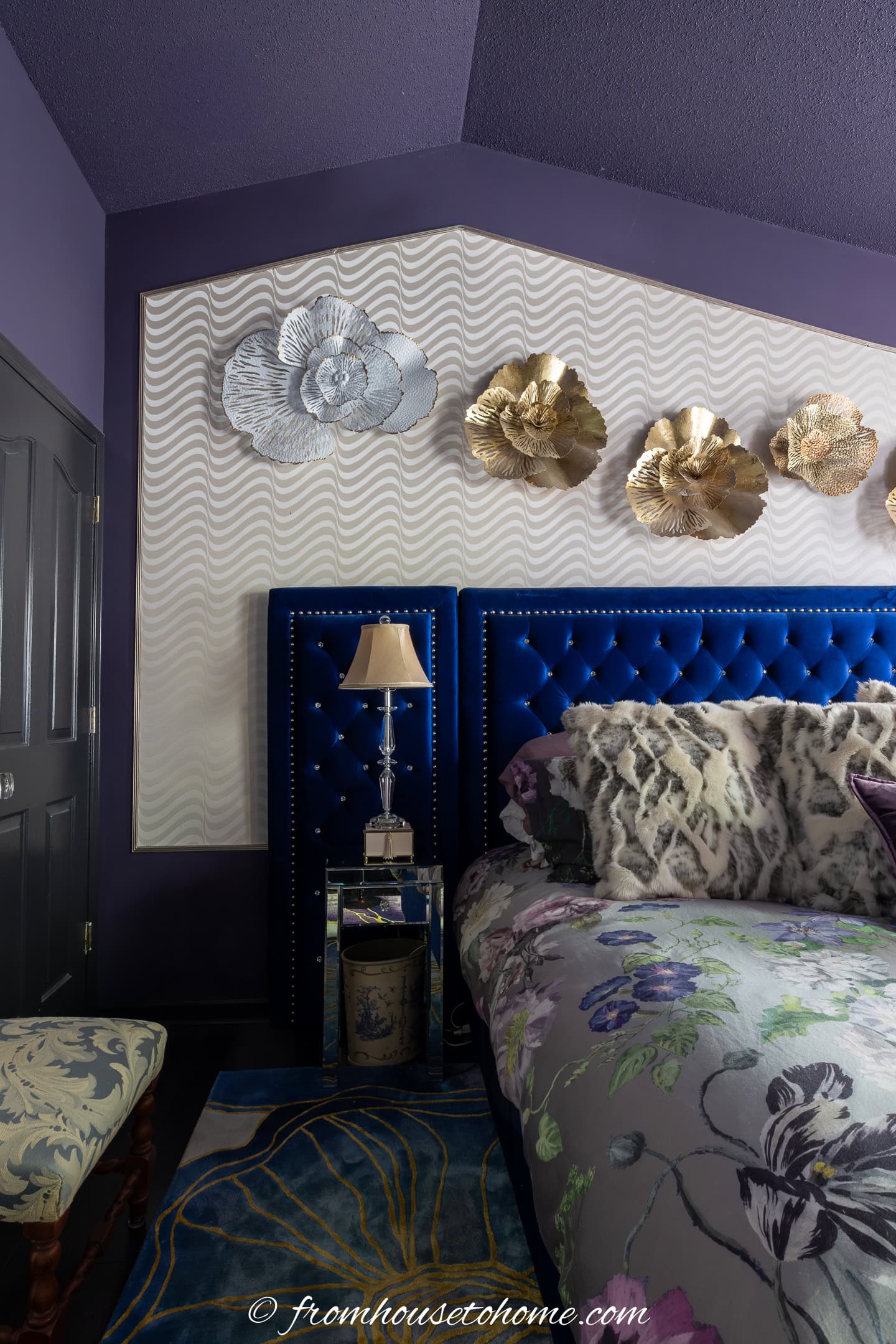 A bedroom with a blue upholstered headboard, floral bedding, metallic wall art, a mirrored nightstand with a lamp, and purple walls.