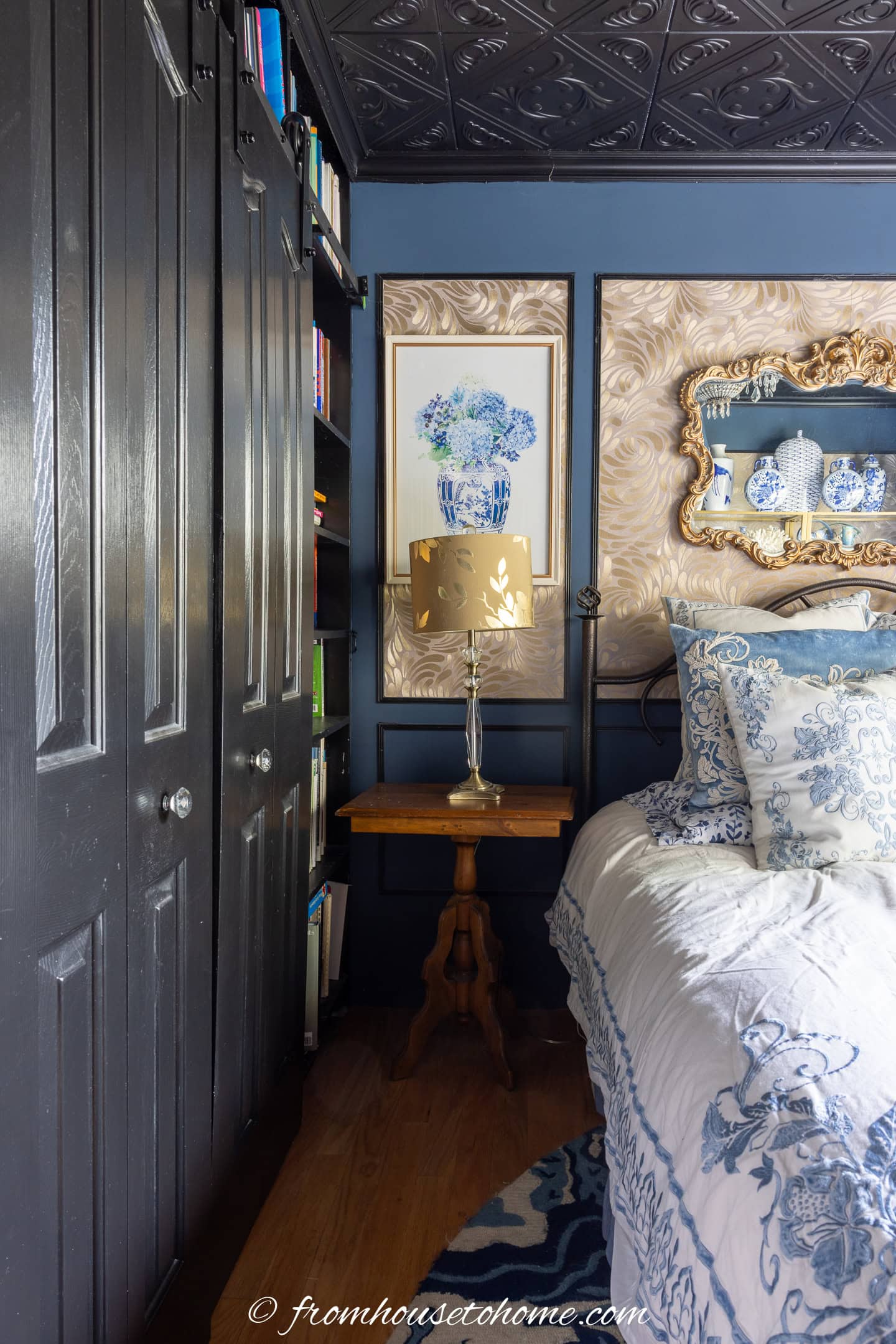 A small bedroom corner with a bed, a wooden nightstand, a gold lamp, and blue floral decor on the wall, next to black closet doors and a bookshelf.