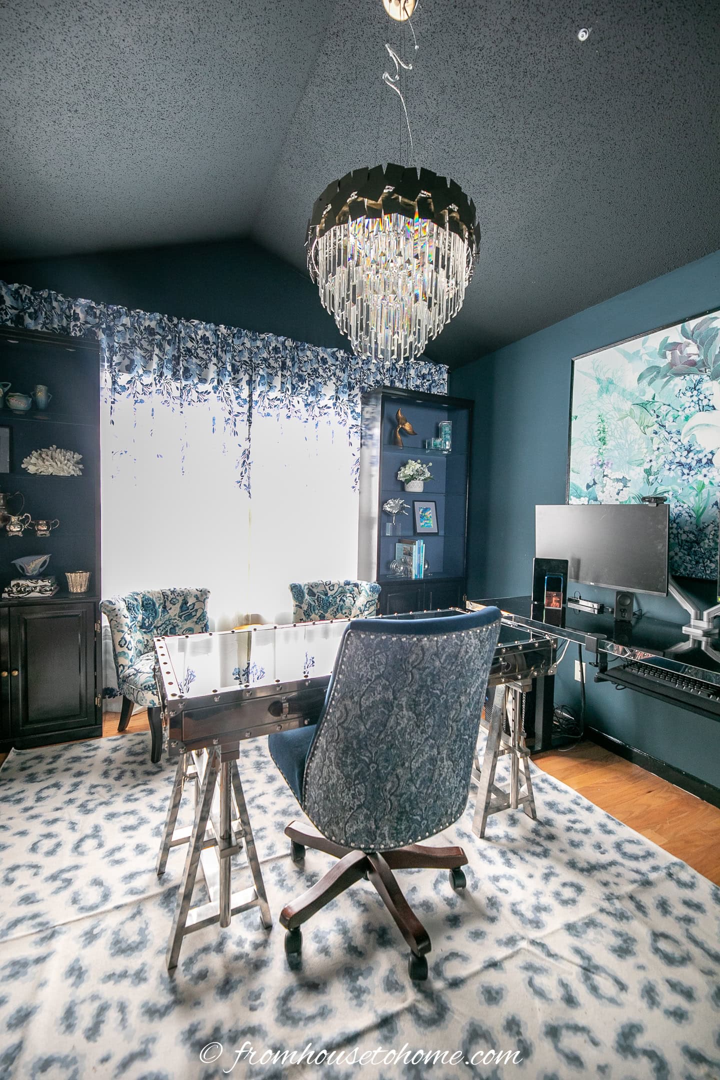 Home office with a patterned chair at a glass desk, shelves filled with decor, a chandelier, computer monitor, and large window with blue and white curtains.
