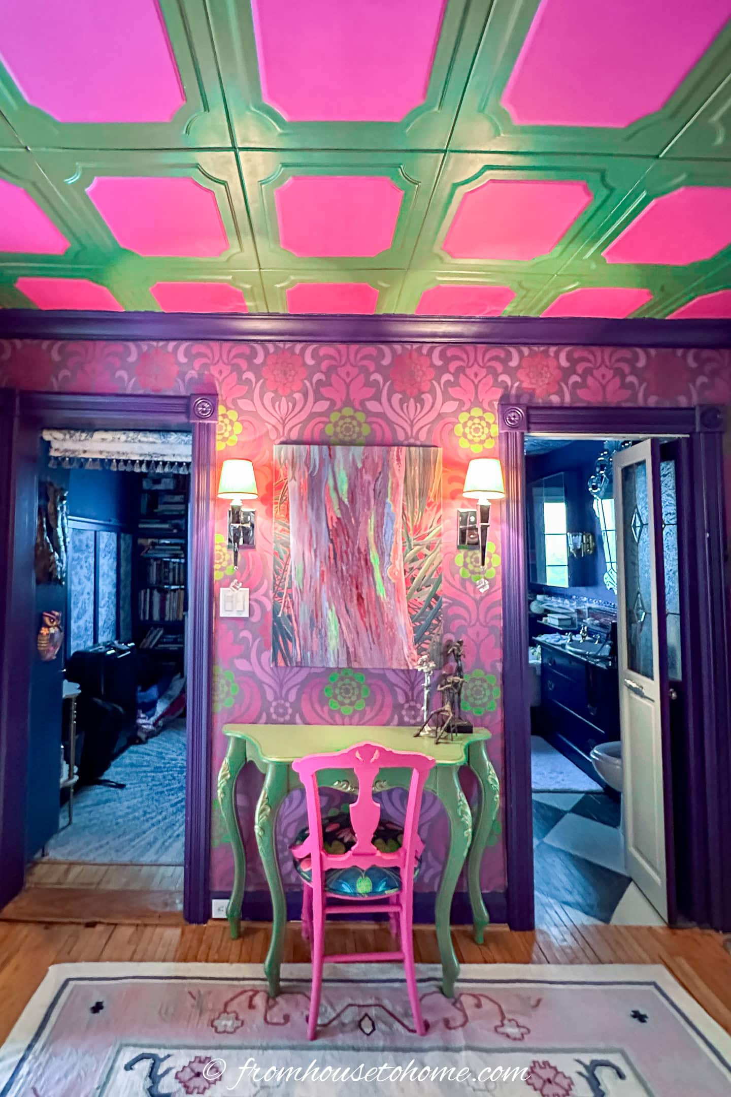 A colorful room with a green and pink coffered ceiling, patterned purple wallpaper, a green table with a pink chair, wall sconces, and a doorway leading to a bathroom.