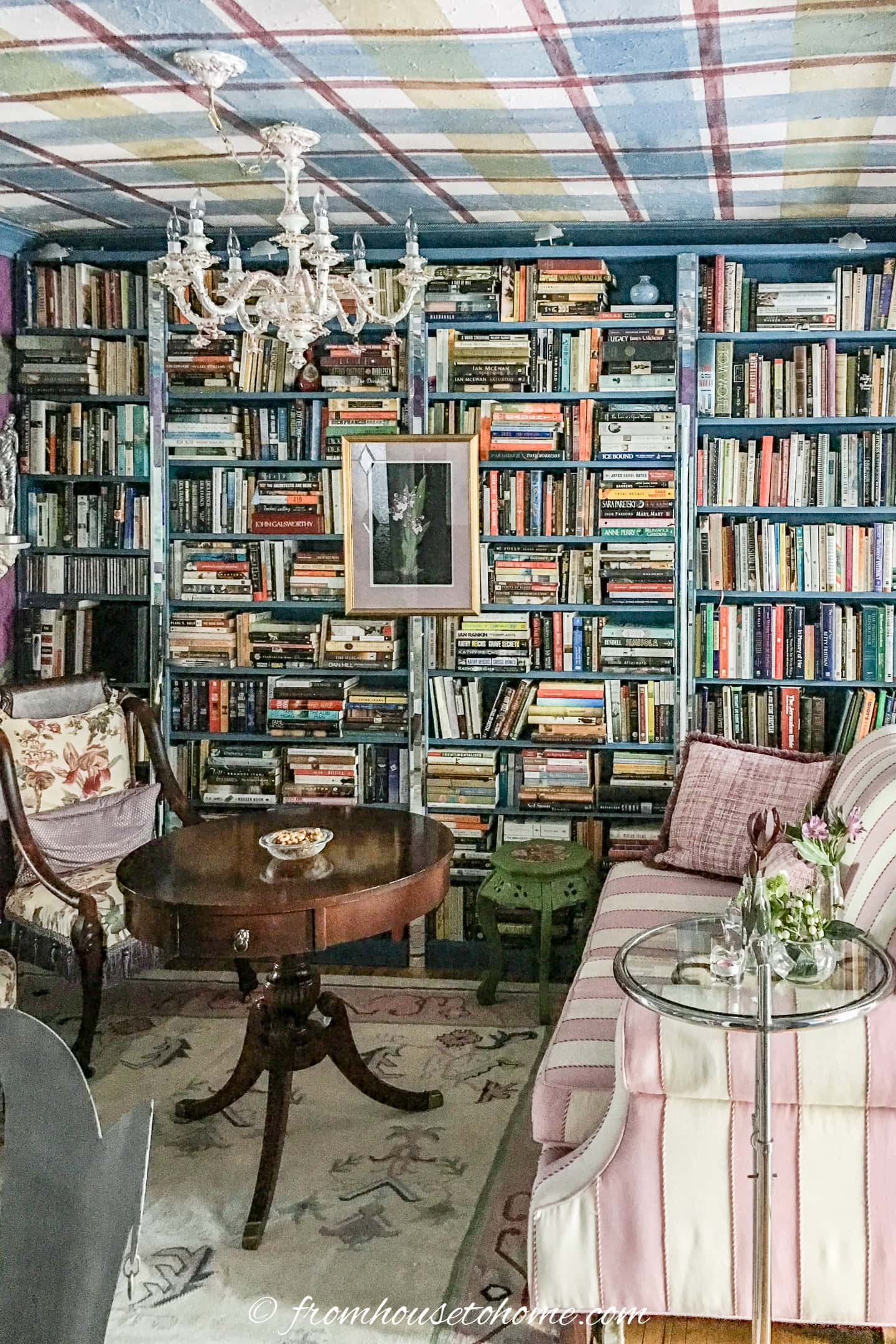 A cozy room with floor-to-ceiling bookshelves, a round wooden table, a chandelier, a striped sofa, and armchair. A framed picture hangs among the books.