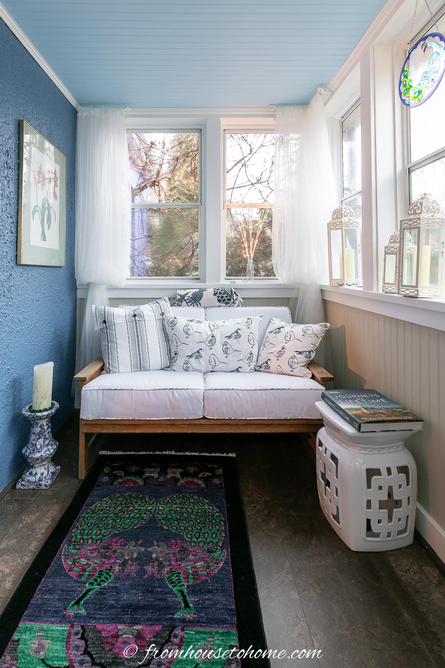 A small sunroom with a white daybed, patterned pillows, a blue accent wall, windows with sheer curtains, a side table with books, and a colorful peacock rug on the floor.