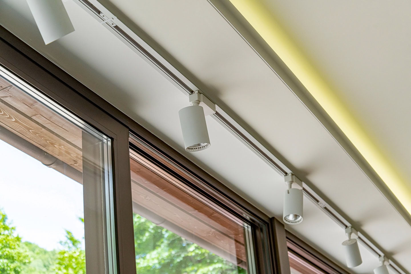 Ceiling-mounted track lighting fixtures above large windows with a view of greenery outside. The track lights are white and modern in design.