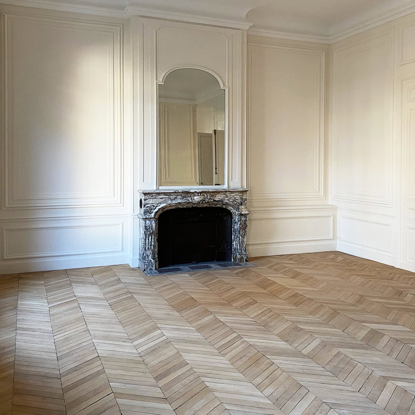 Empty room with herringbone wood floors, cream-colored paneled walls, a marble fireplace, and a large arched mirror above the mantel.
